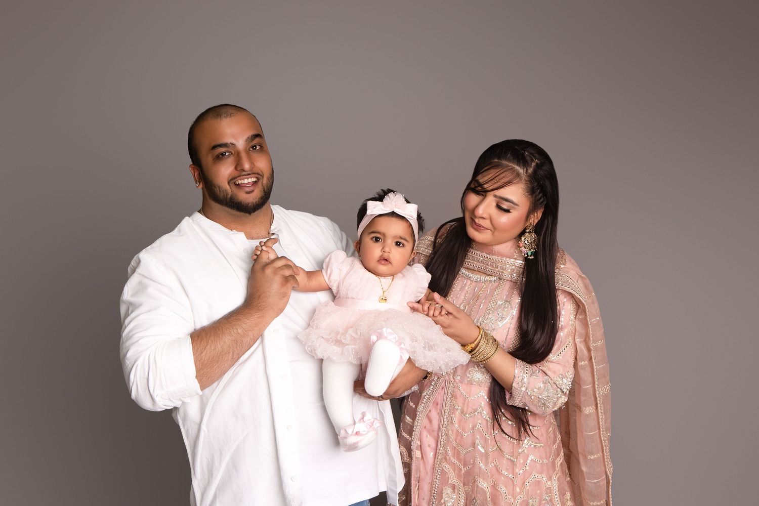 A family portrait of a man, woman, and baby girl in pink, posing together against a gray studio background.