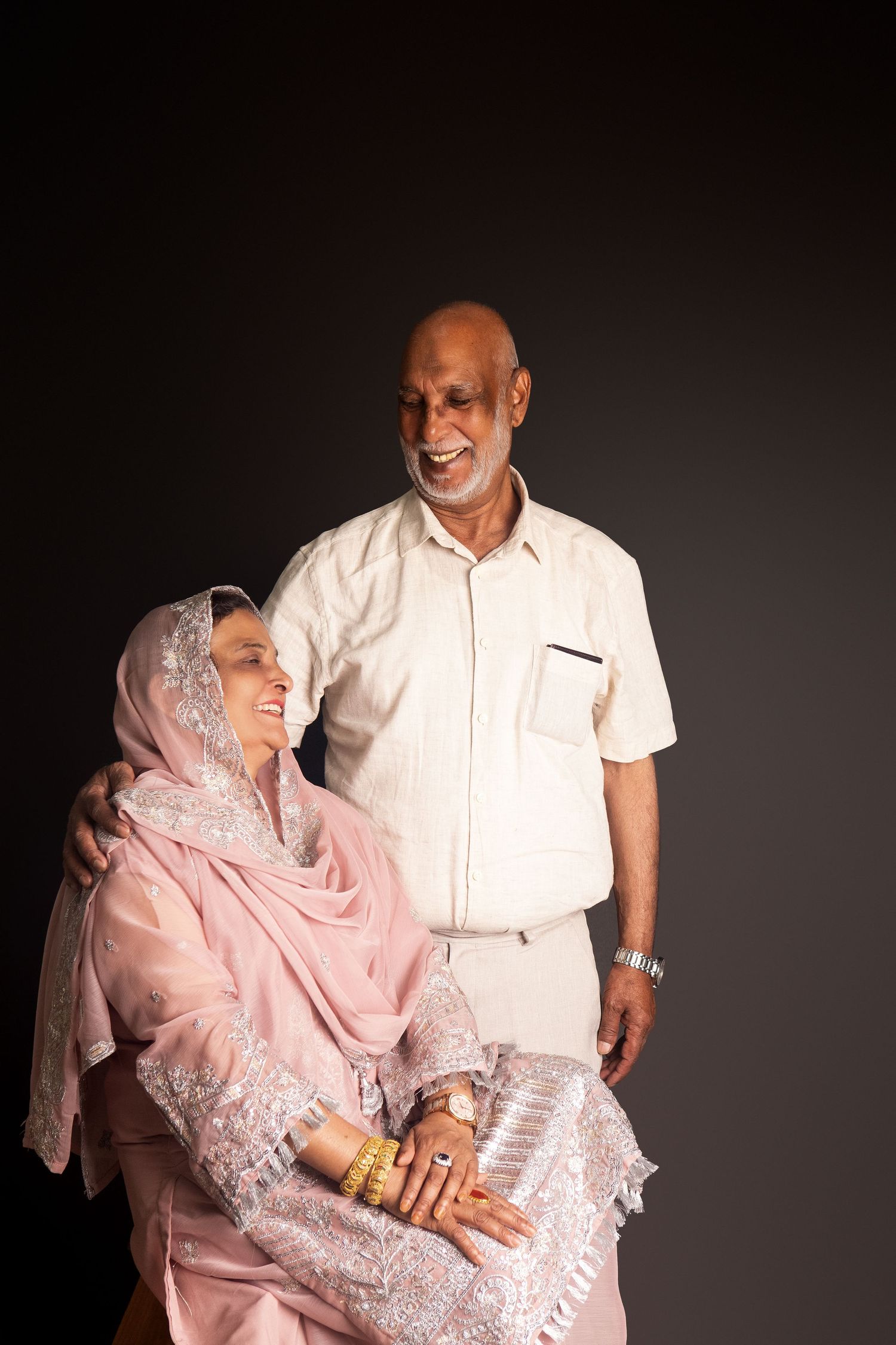 Elderly Indian couple in traditional attire, woman in pink salwar seated, man in white shirt standing, smiling warmly.