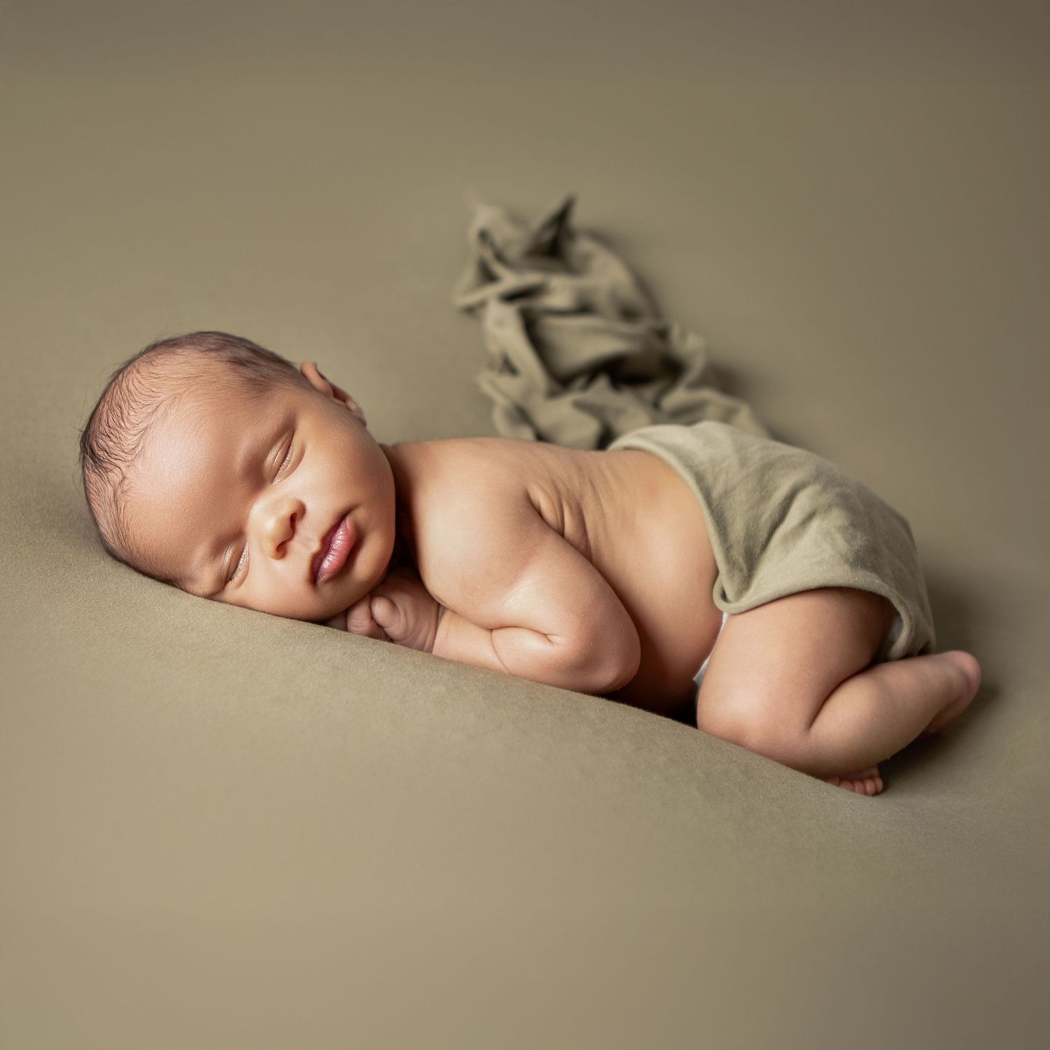 Peaceful newborn baby sleeping curled up on olive green backdrop wearing matching wrap pants in professional studio photo.