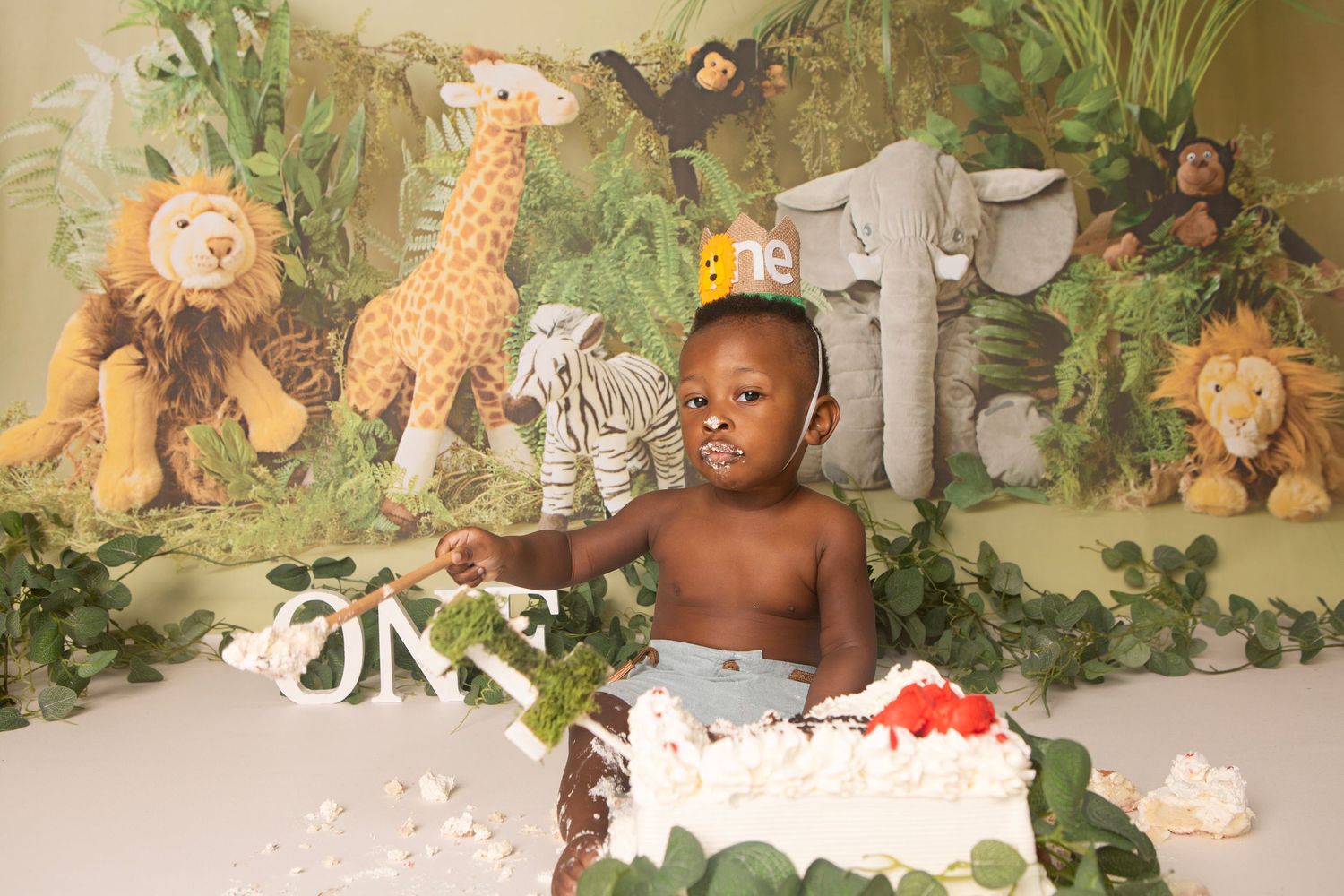 Baby boy celebrates first birthday with jungle-themed cake smash surrounded by stuffed safari animals and tropical backdrop.