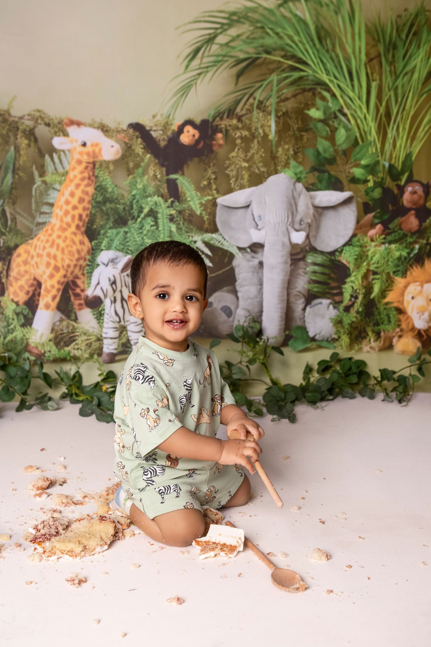 Smiling baby boy in jungle romper sits on floor with spoon amid cake smash, safari animal backdrop behind him.