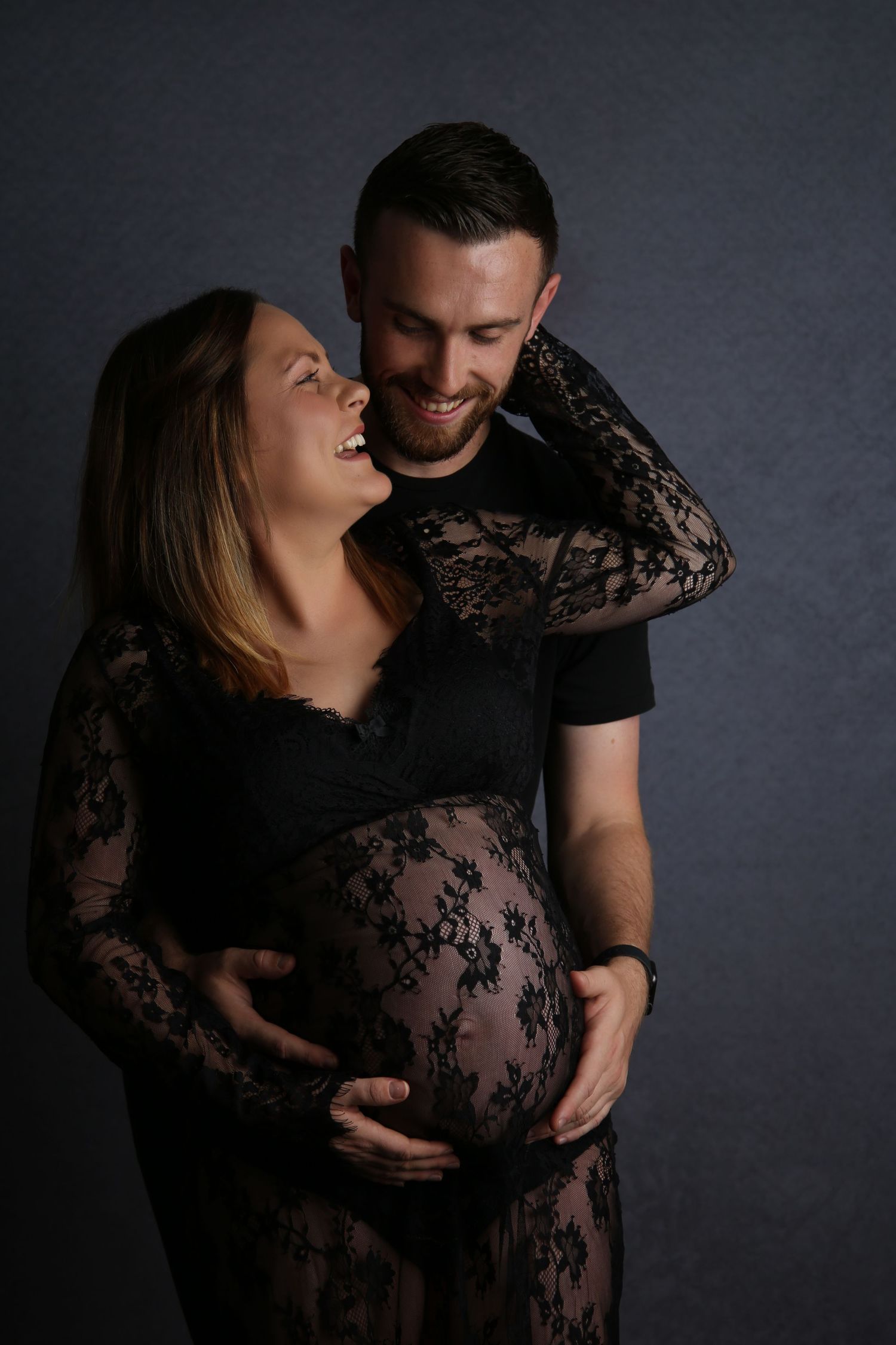 Smiling couple in black outfits cradling pregnant belly during maternity photo shoot against dark background.