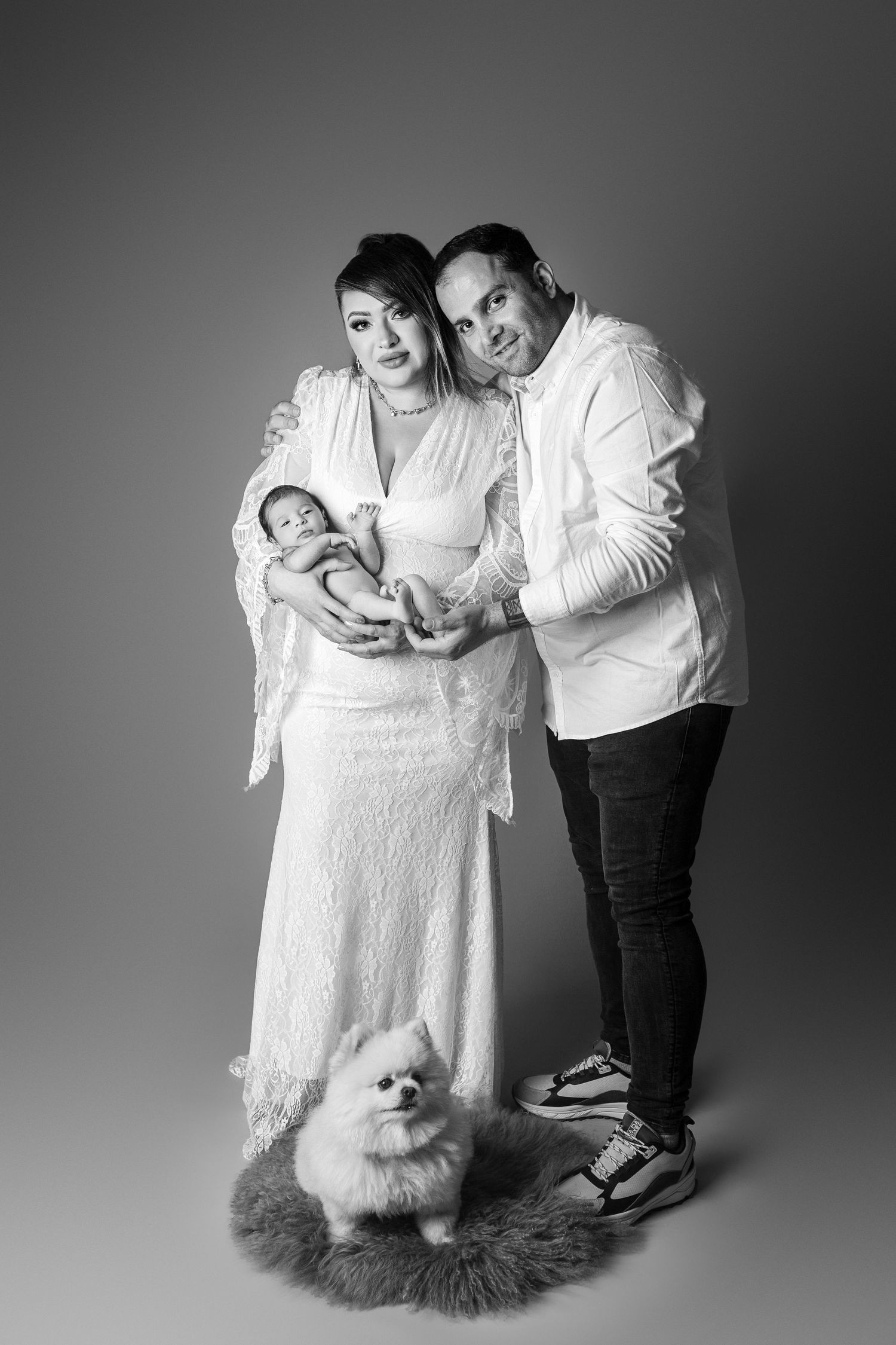 Black and white family portrait with couple holding newborn baby and small Pomeranian dog on fur rug in studio.