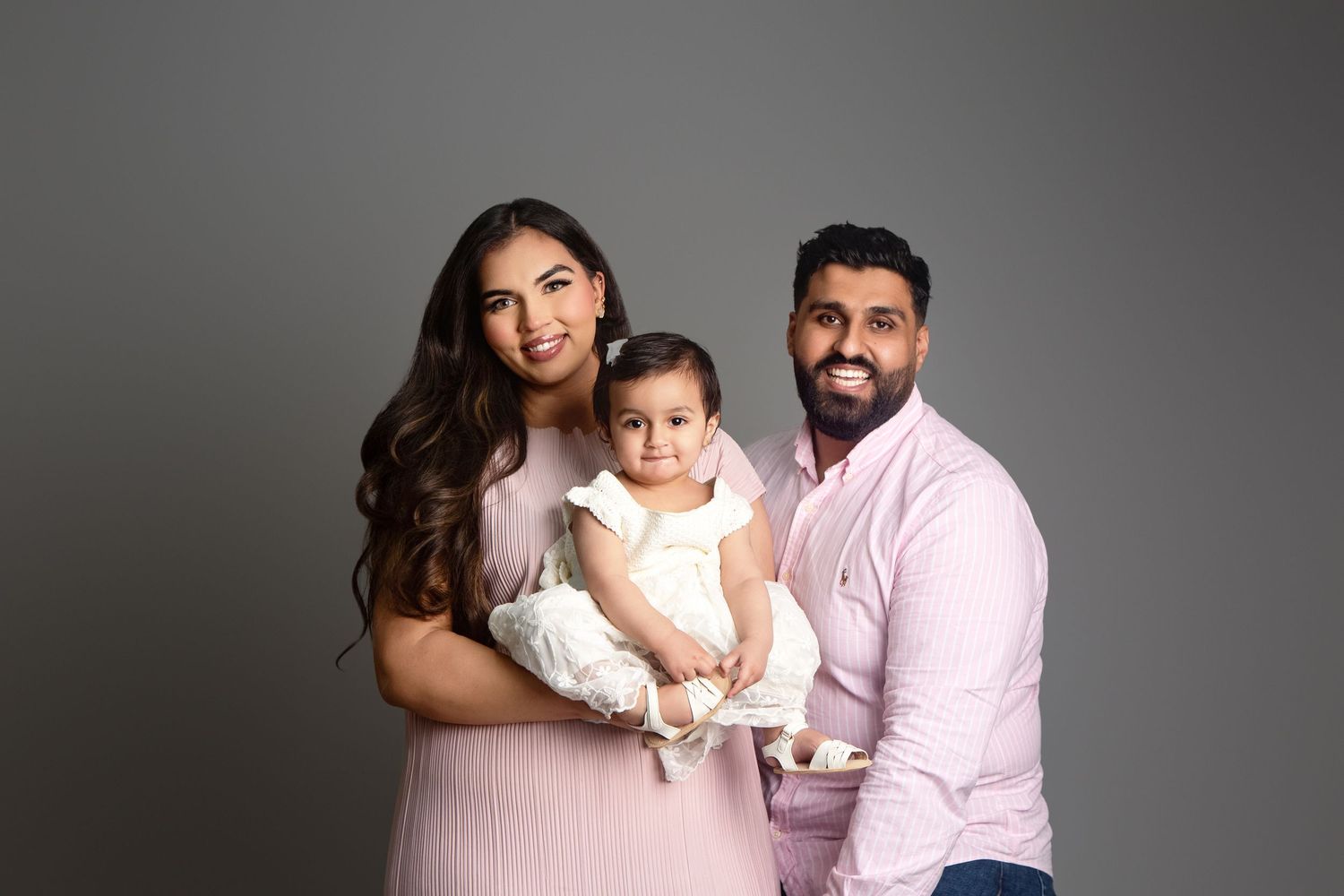 Happy family portrait with parents holding baby girl in white dress, studio photo on gray background.