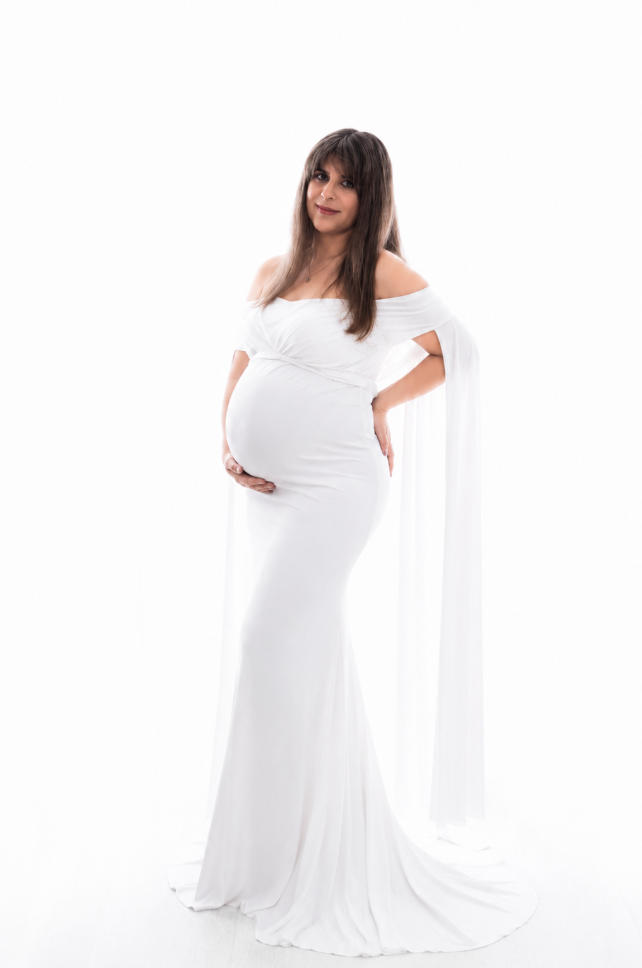Pregnant woman posing confidently in fitted white off-shoulder maternity gown with draped sleeves.