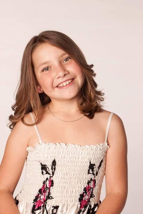 Smiling young girl with brown hair wearing a floral sundress poses for a portrait photo against a light background.