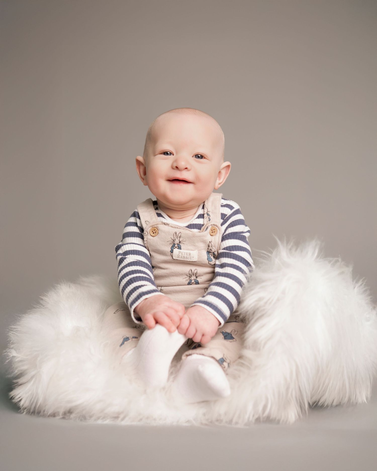 Smiling baby in striped shirt and overalls sitting on fluffy white fur rug against gray background.