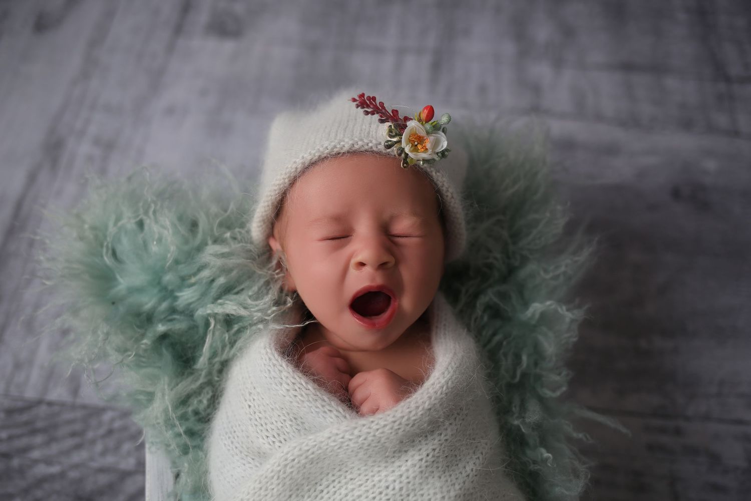 Newborn baby yawning wrapped in white knit blanket wearing floral hat on fluffy mint green fur backdrop.