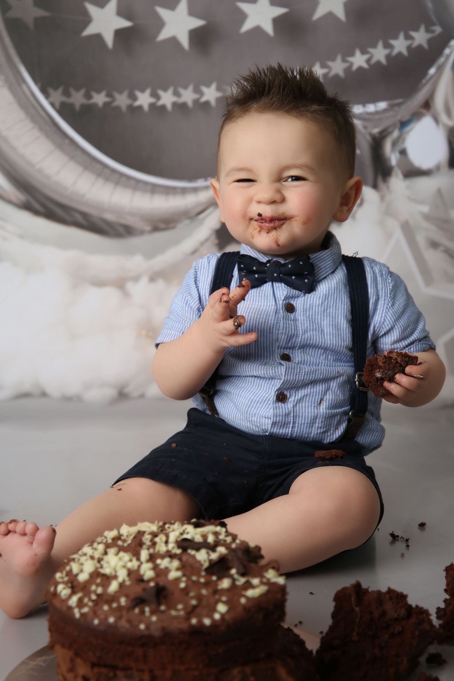 Adorable baby boy in bow tie and suspenders smashes chocolate birthday cake, face covered in frosting.