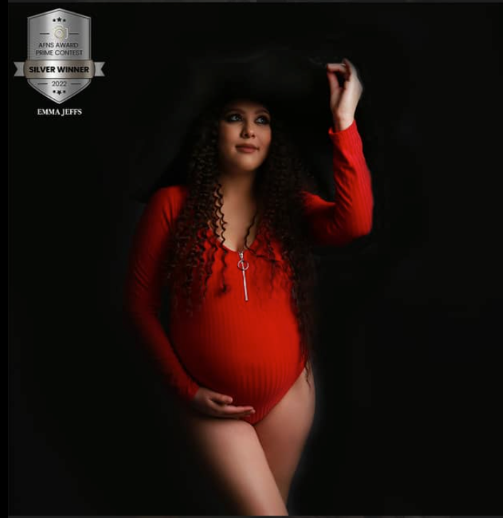 A pregnant woman in a red bodysuit and black hat poses elegantly in a dark studio maternity portrait.