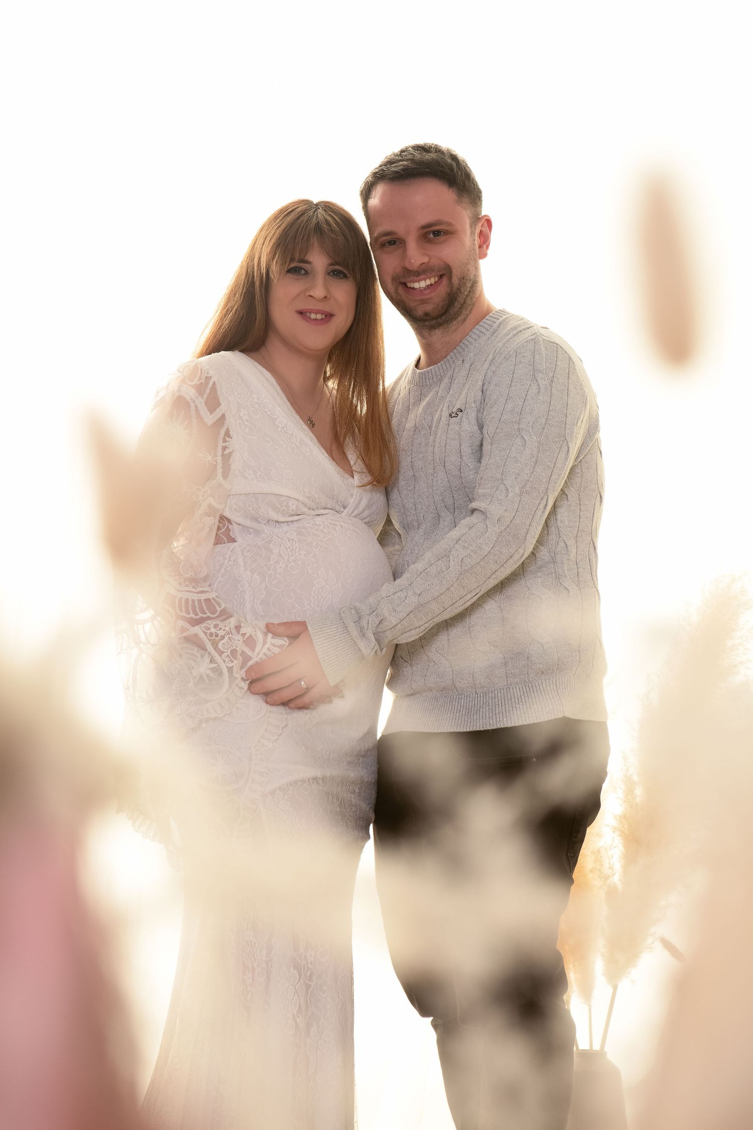 Couple posing in sunlit field, pregnant woman in white lace dress embraced by smiling partner.