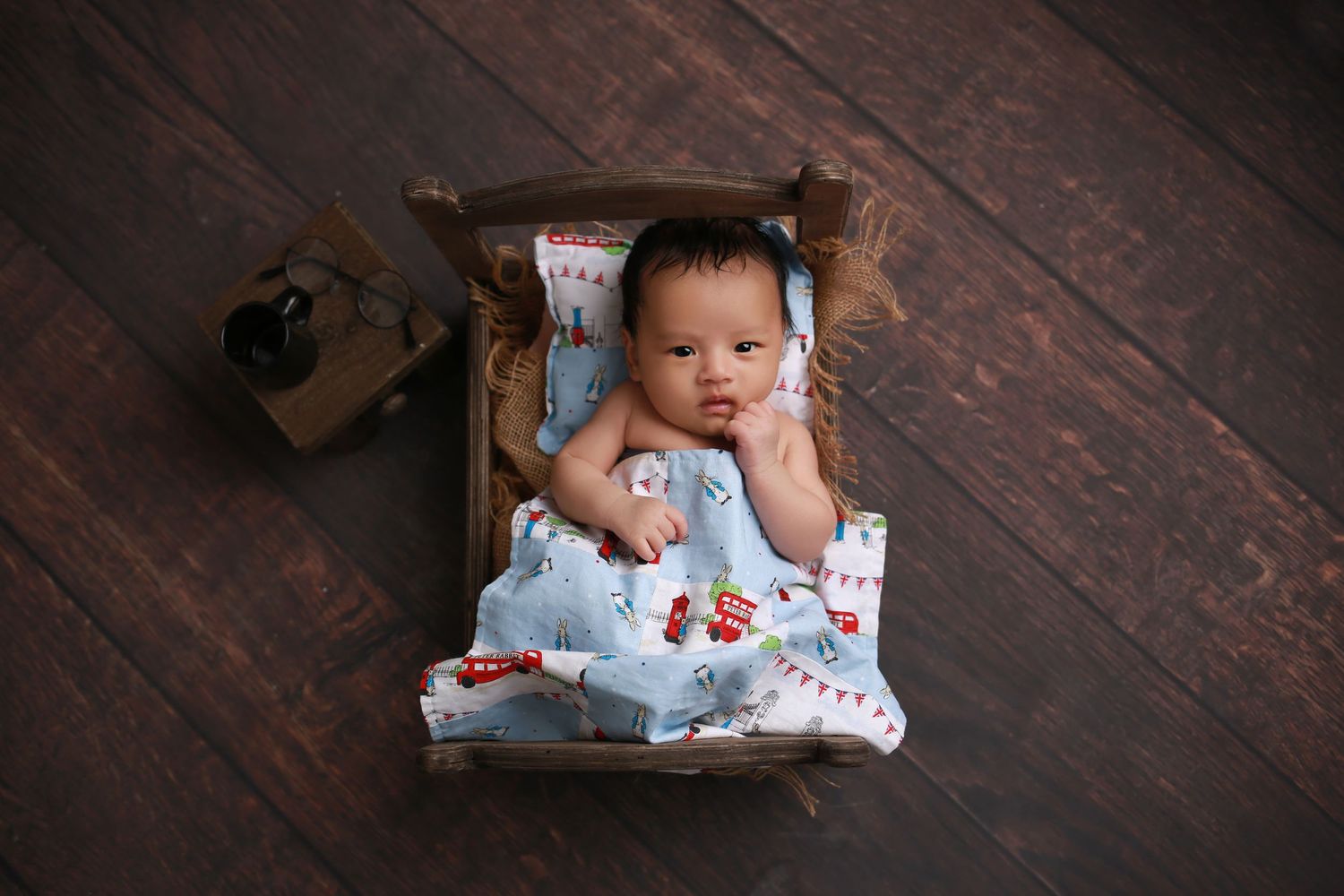 Newborn baby lying in a small wooden bed wrapped in a light blue Christmas-themed blanket on a dark wood floor.