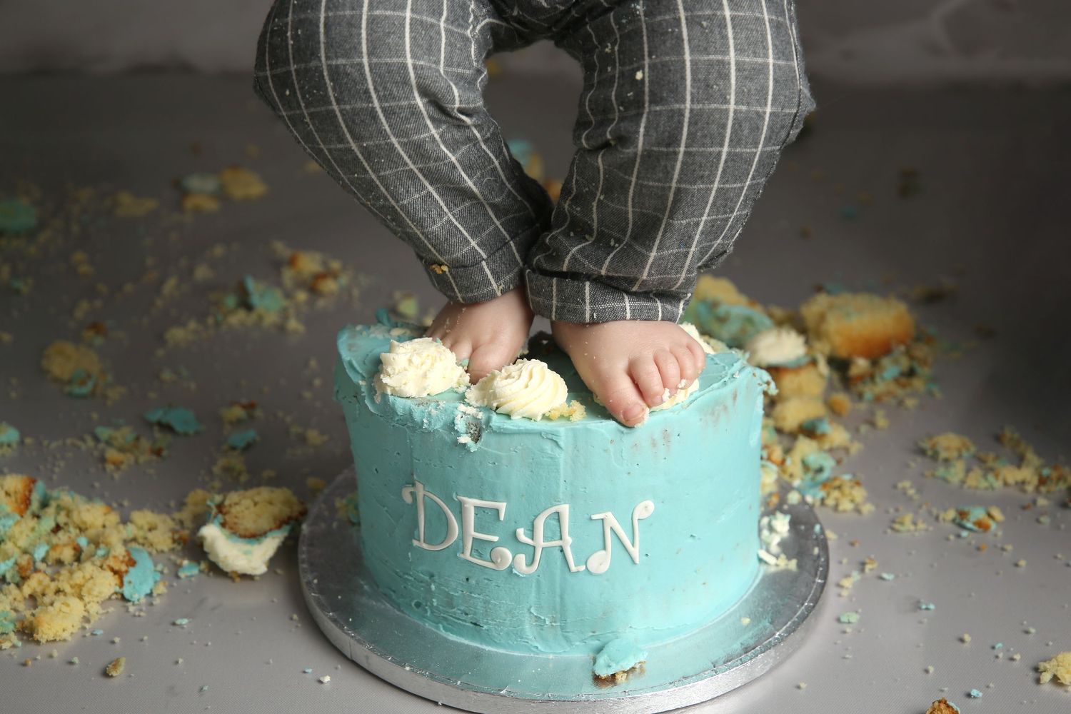 Baby feet smashing a teal first birthday cake with the name Dean written in white frosting.