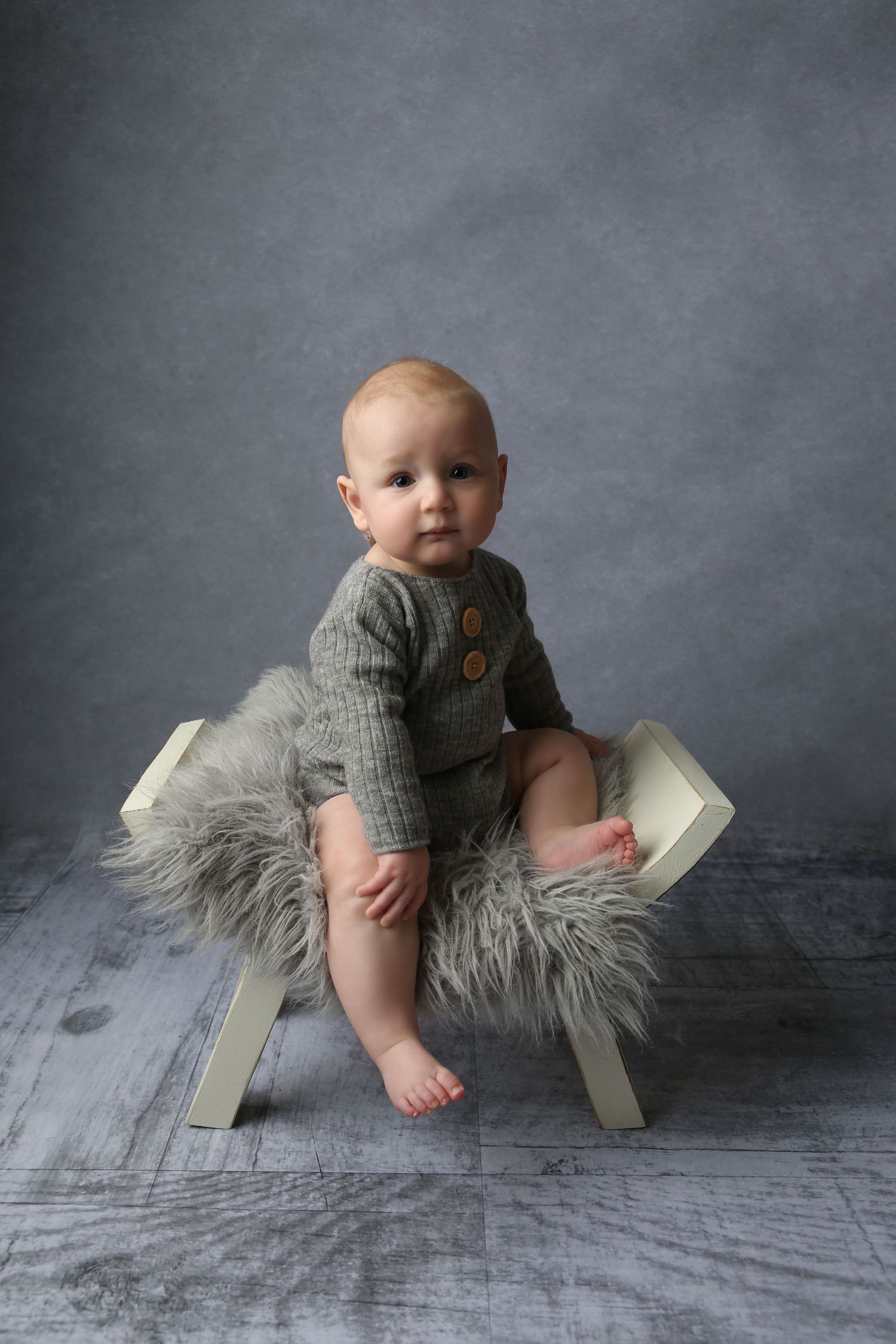 A baby in a gray knit romper sits on a small white bench with fluffy fur, posed against a gray backdrop.
