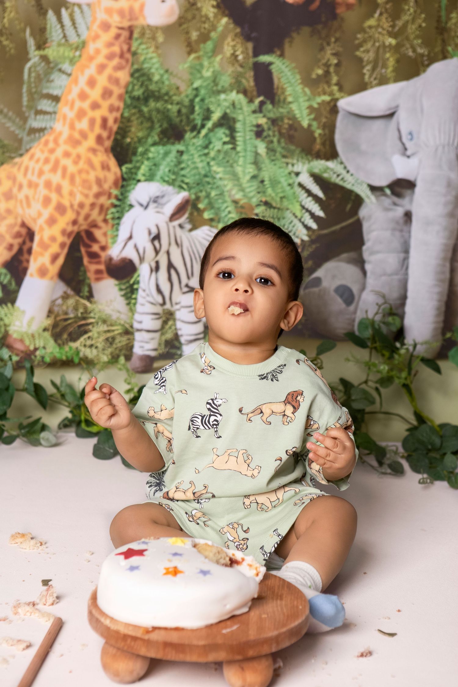 Baby boy in safari onesie sits before smash cake, with jungle animal backdrop featuring giraffe and elephant.