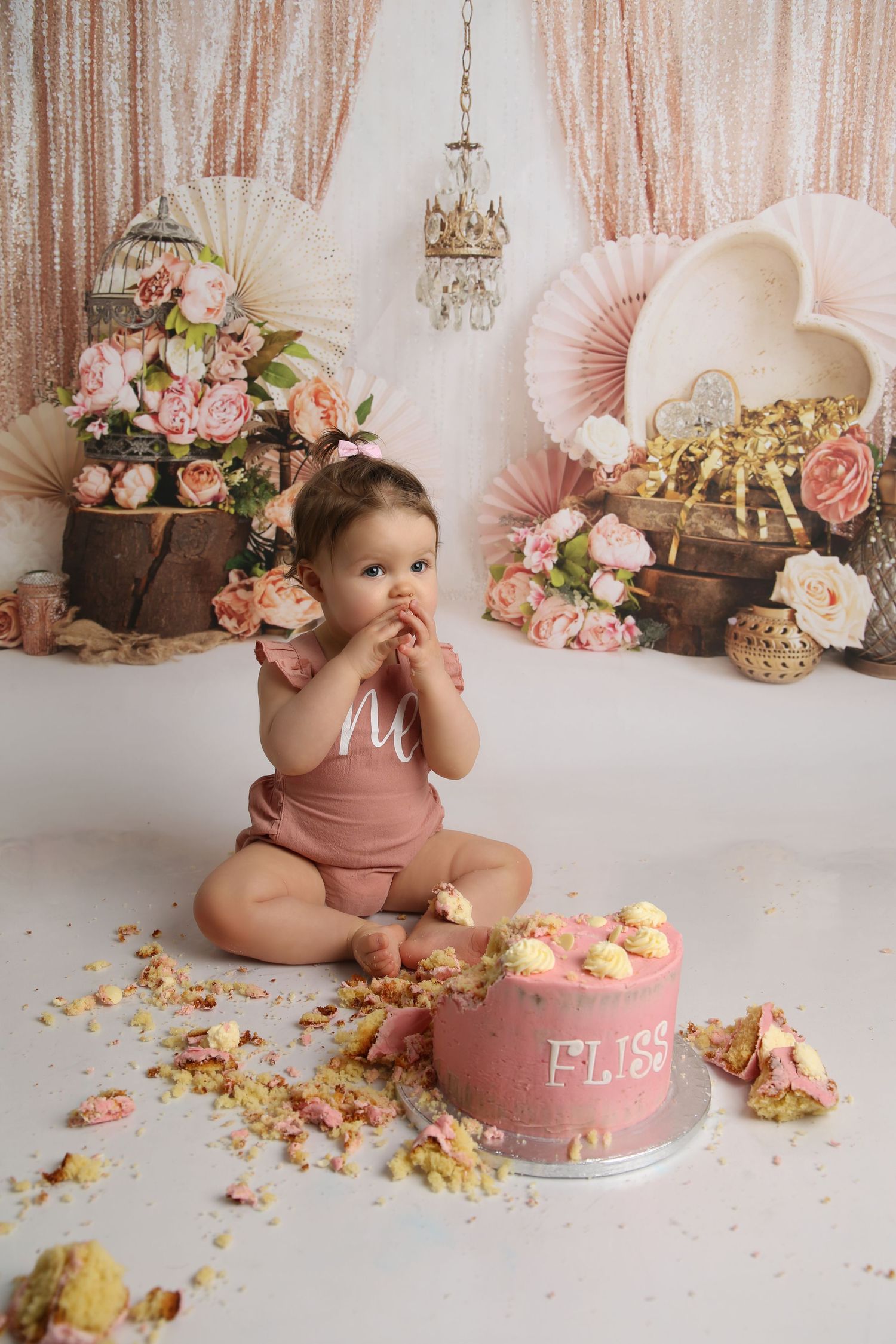 Baby girl in pink romper enjoying her first birthday cake smash with floral decorations and a pink cake reading Fliss.