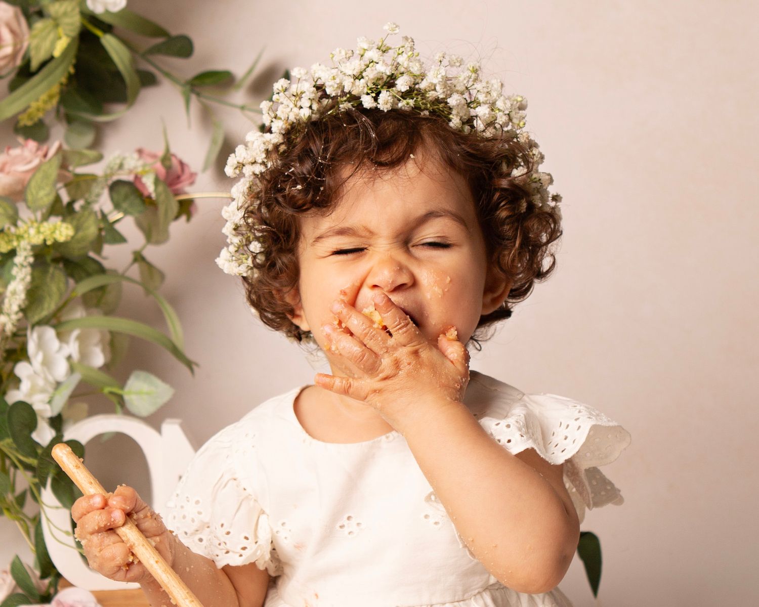 Toddler girl in white dress and floral crown joyfully eating cake with her hands during a cake smash photoshoot.