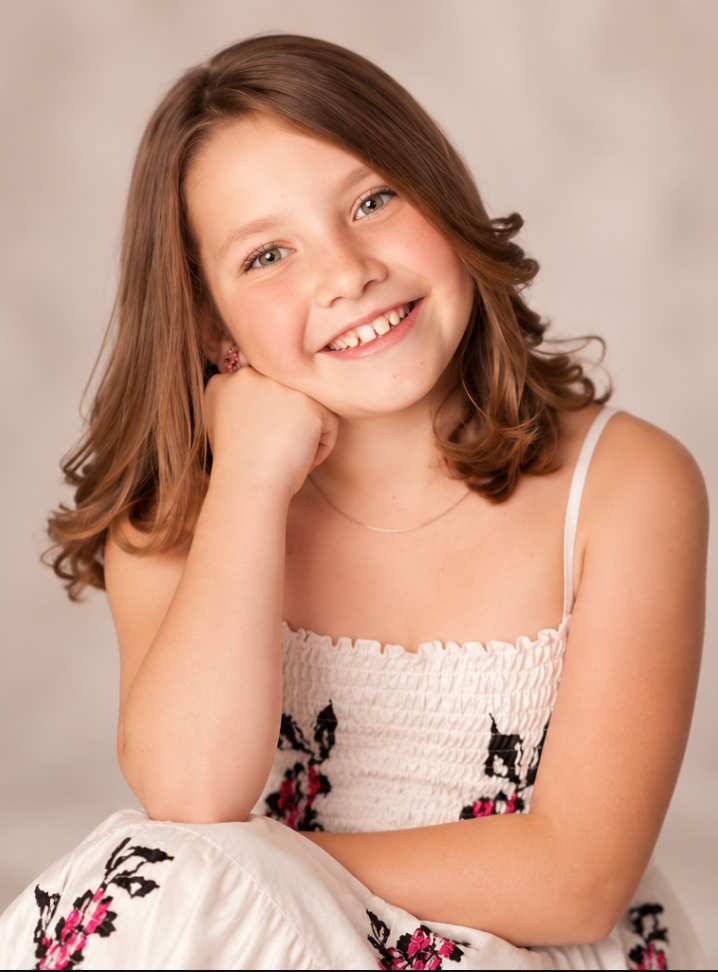 Smiling young girl with brown hair wearing a floral white dress, posing for a portrait photo.