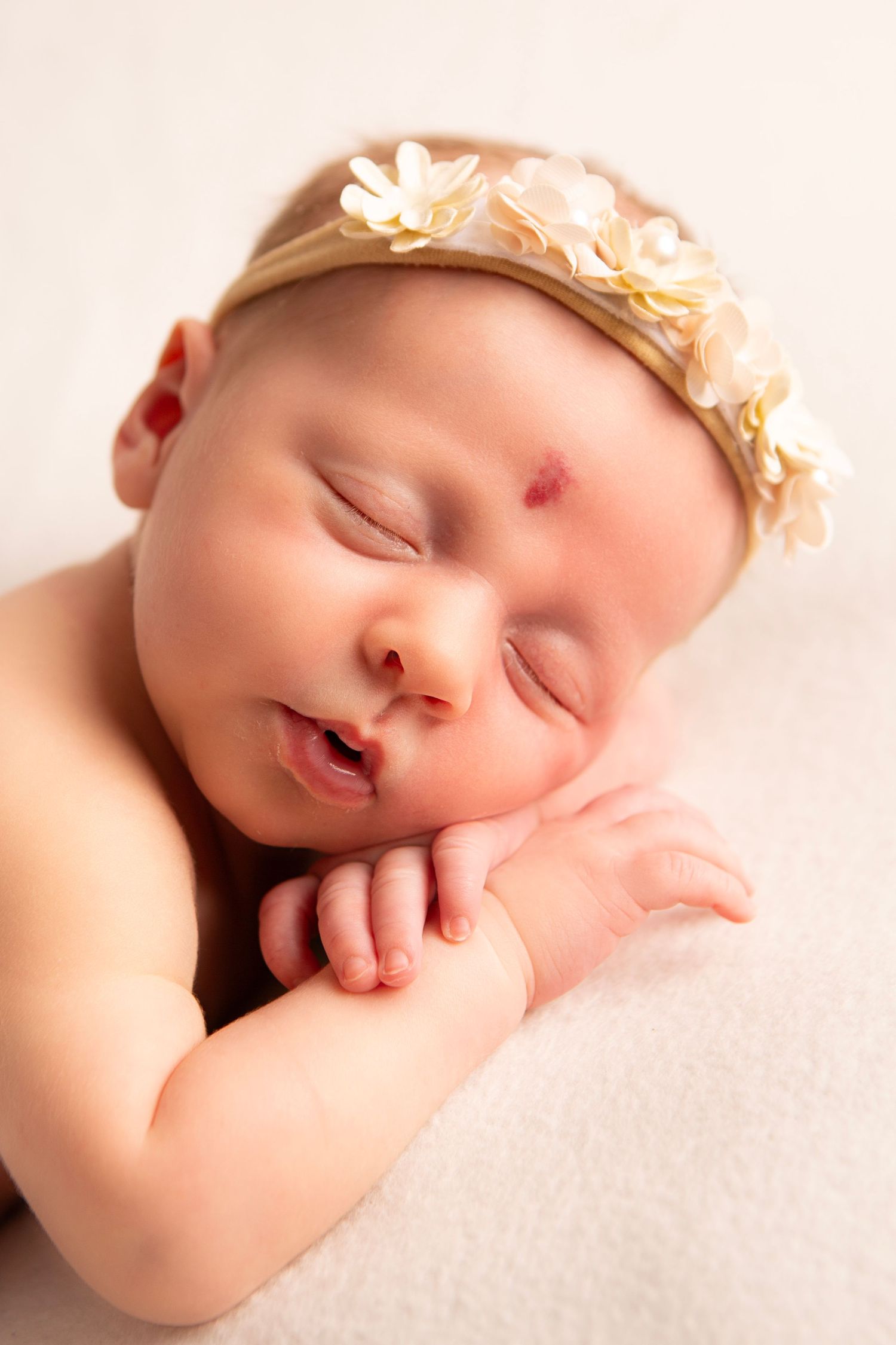Sleeping newborn baby girl wearing a delicate floral headband, resting peacefully with hands tucked under her chin.