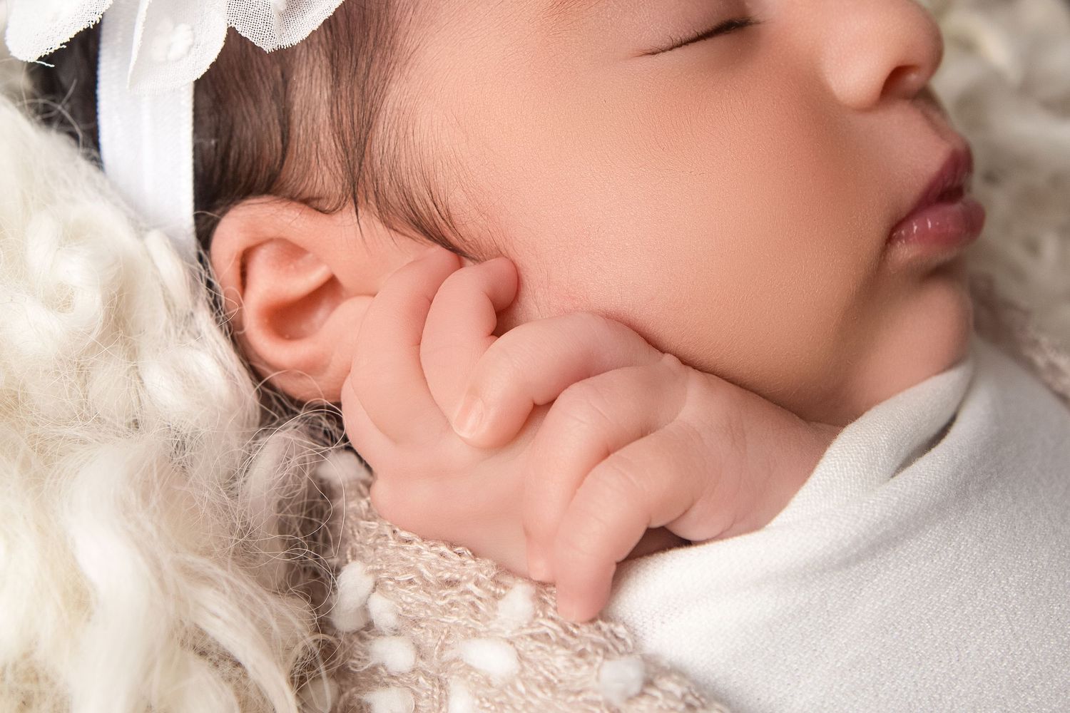 Close-up of a sleeping newborn baby with tiny hand near face, wearing white bow headband, nestled in soft woolly blanket.