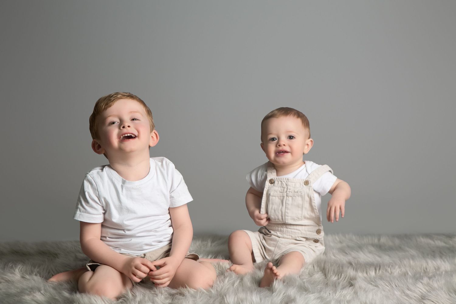 Two happy baby boys sitting on a fluffy gray rug, wearing white onesies, smiling against a gray background.