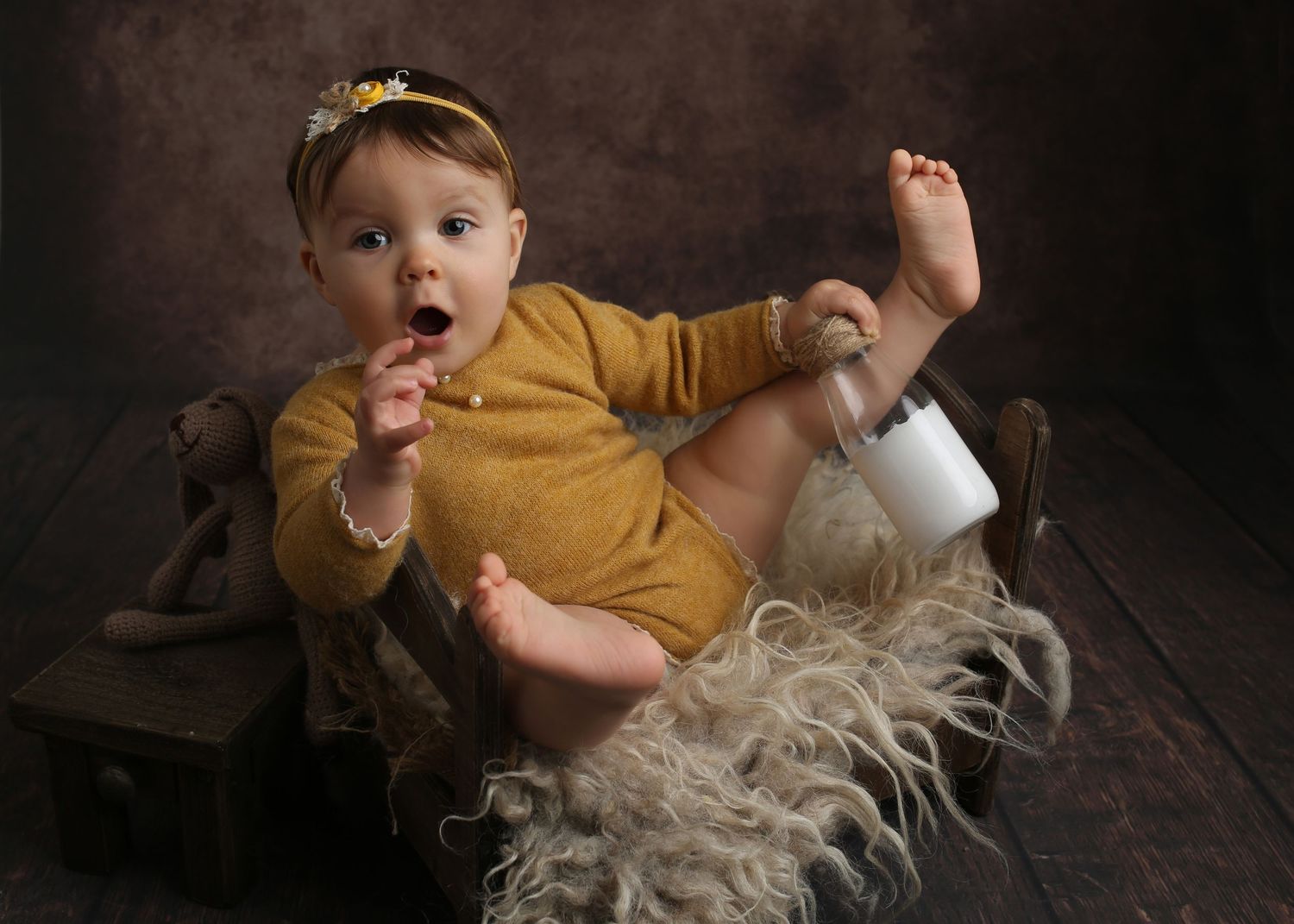 Adorable baby girl in mustard onesie and floral headband sitting in rustic wooden box with fluffy fur.