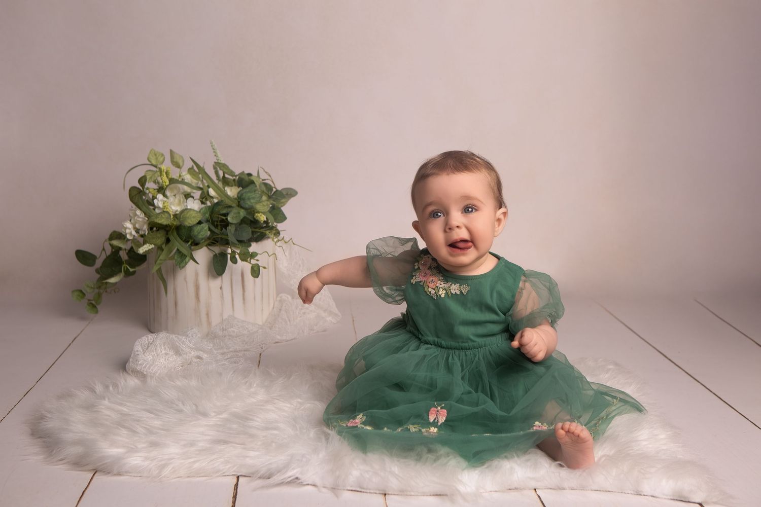 A smiling baby girl in a green tulle dress sits on a white fur rug beside a floral arrangement in a studio.