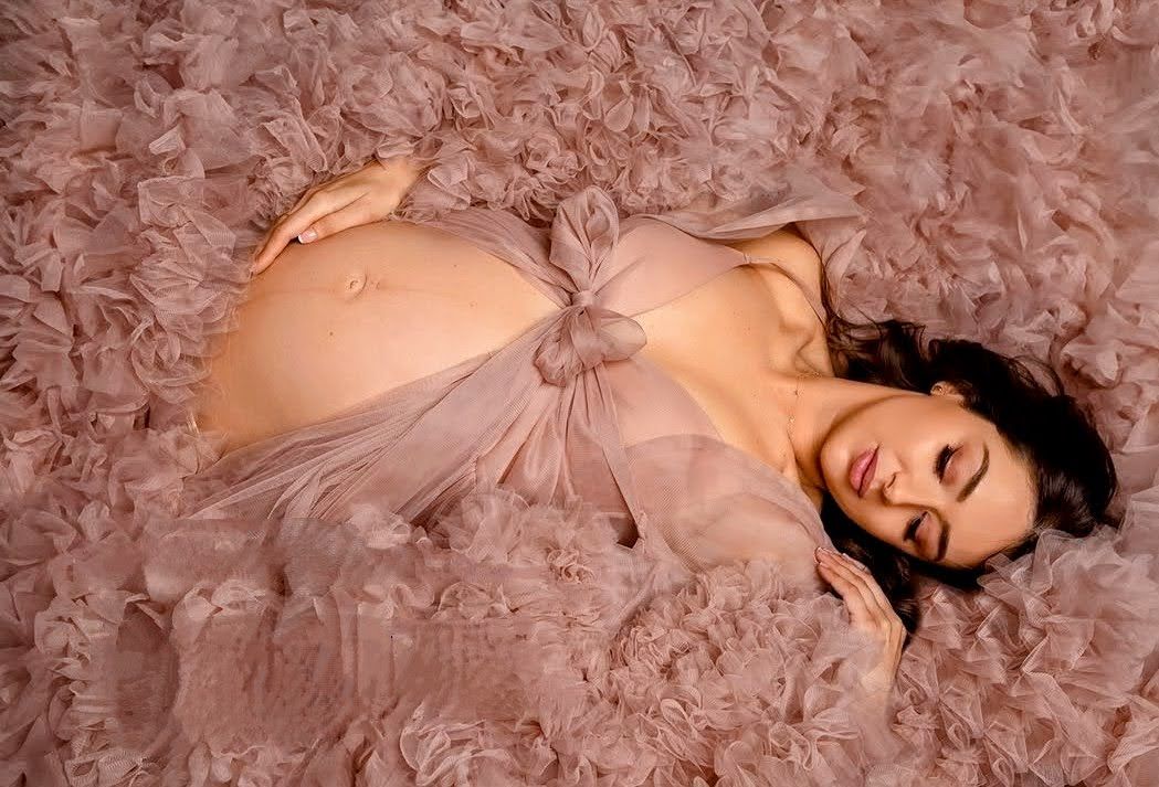 Pregnant woman lying on pink fluffy surface wearing blush tulle dress, showcasing baby bump.