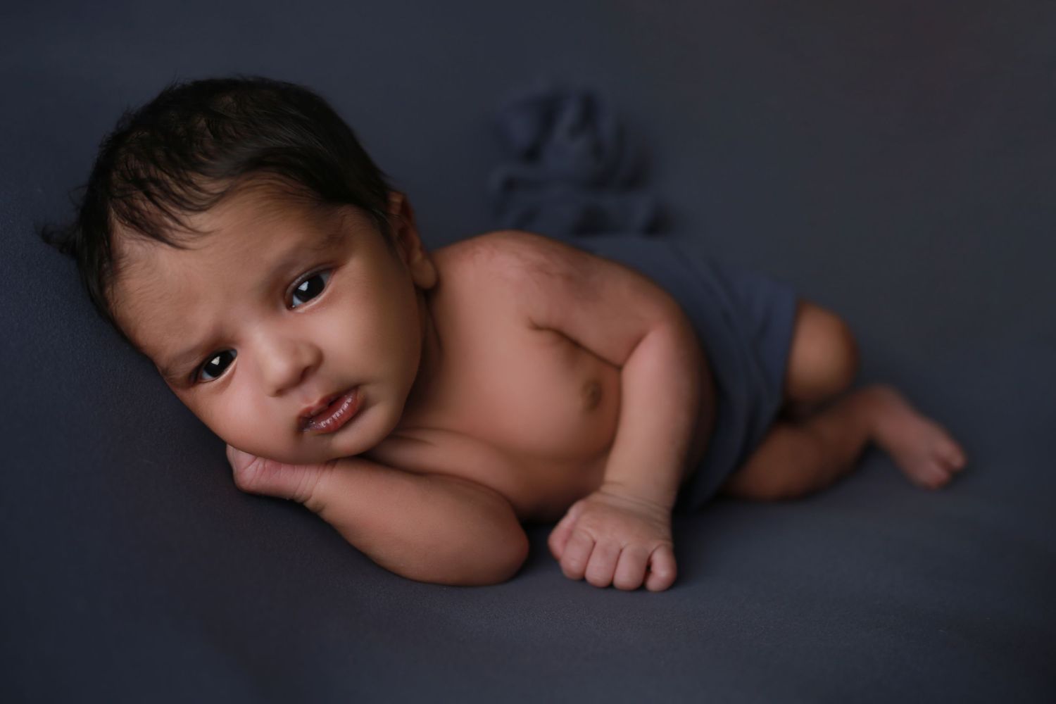 Newborn baby lying on a gray blanket, looking at the camera during a professional newborn photography session.