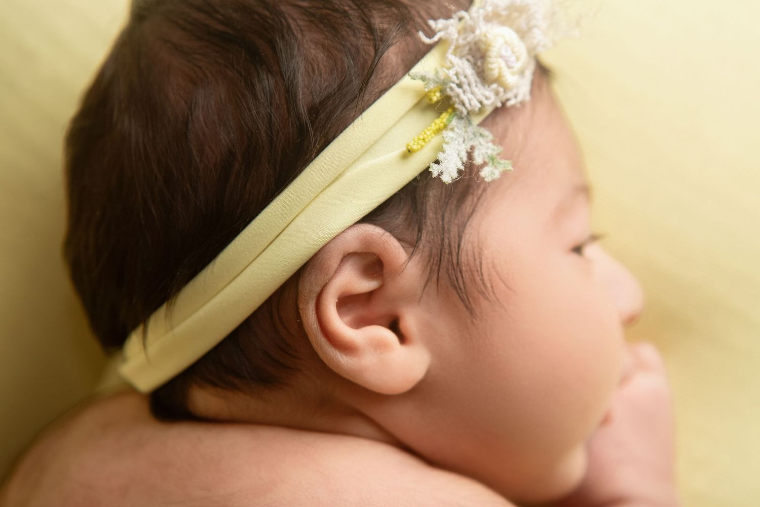 Close-up profile of a newborn baby wearing a delicate yellow floral headband, resting peacefully on a soft green background.