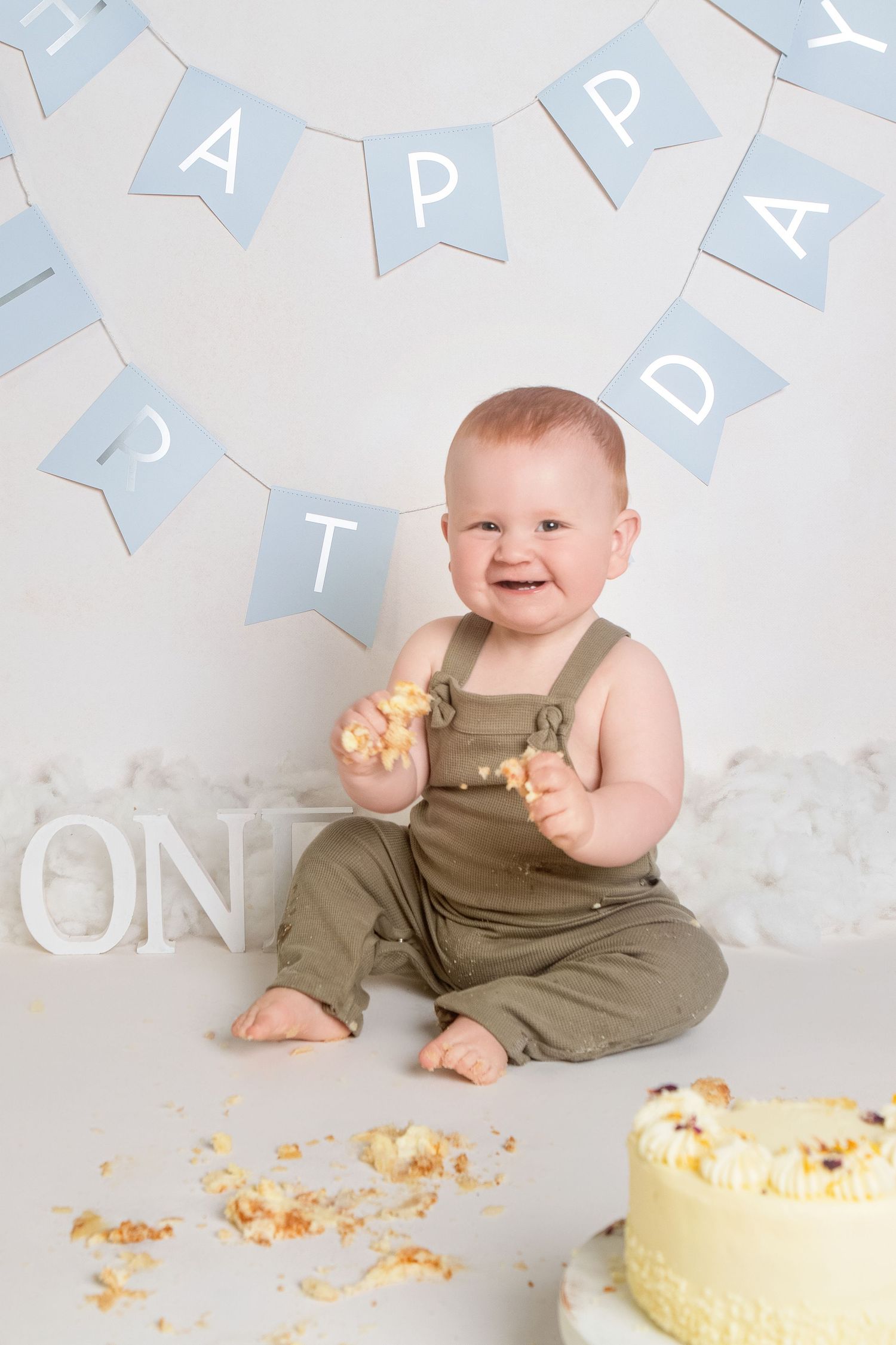 Happy baby in green overalls smashes first birthday cake, sitting in front of blue Happy Birthday bunting.