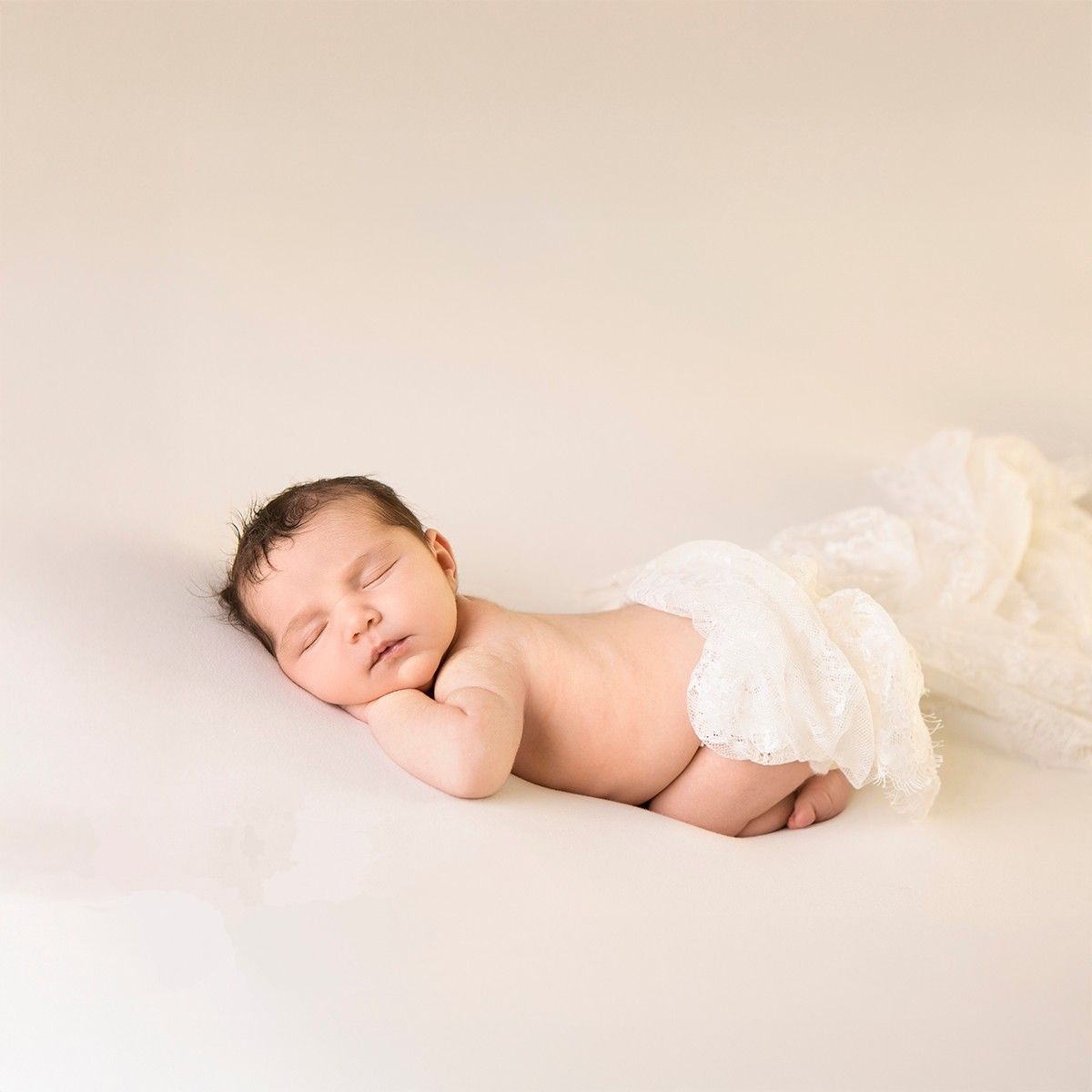 Peaceful newborn baby sleeping on a cream background, wearing a white ruffled diaper cover, in a professional newborn photo.