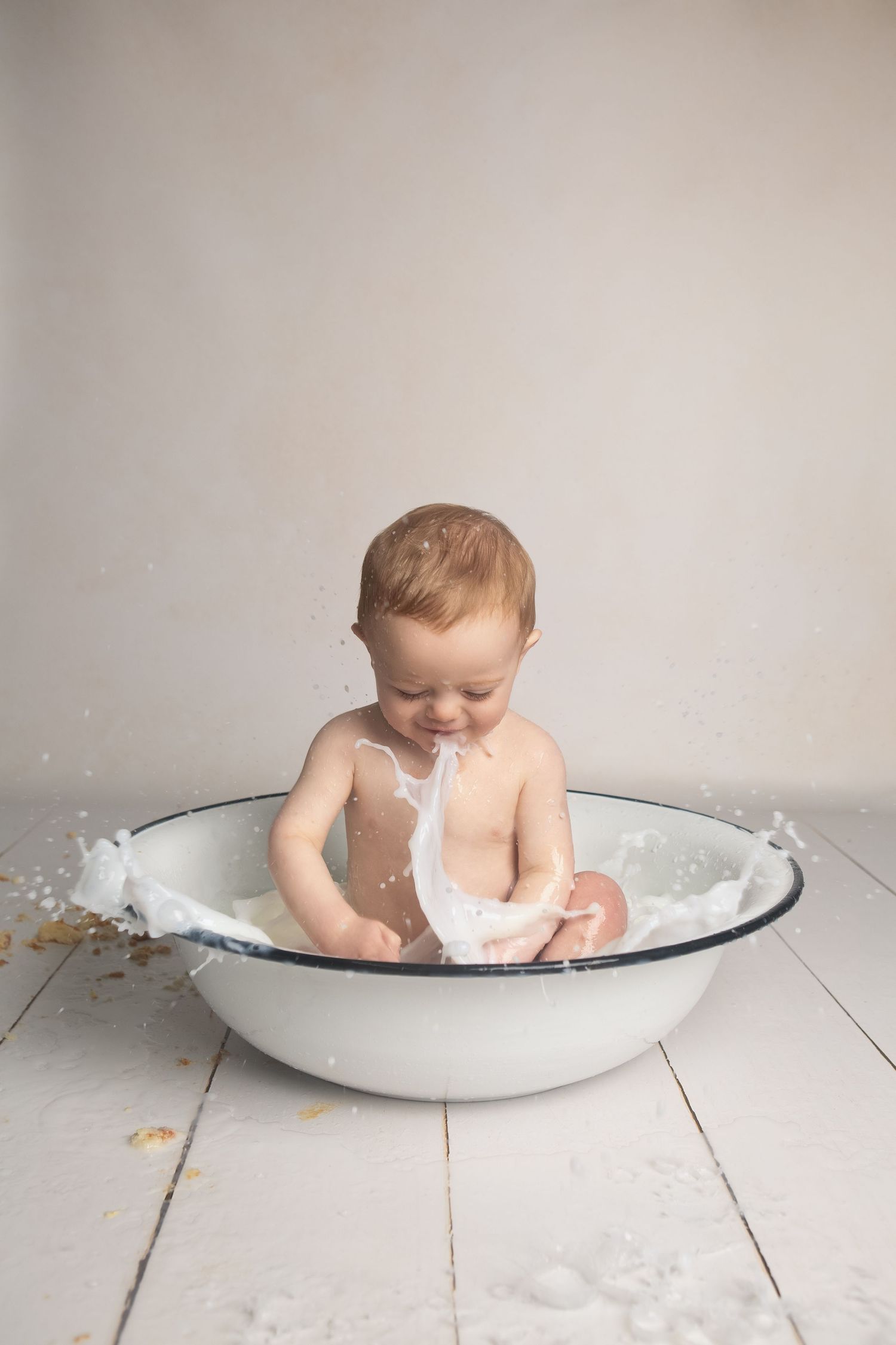 A smiling baby splashing in a white enamel basin tub on a white wooden floor.