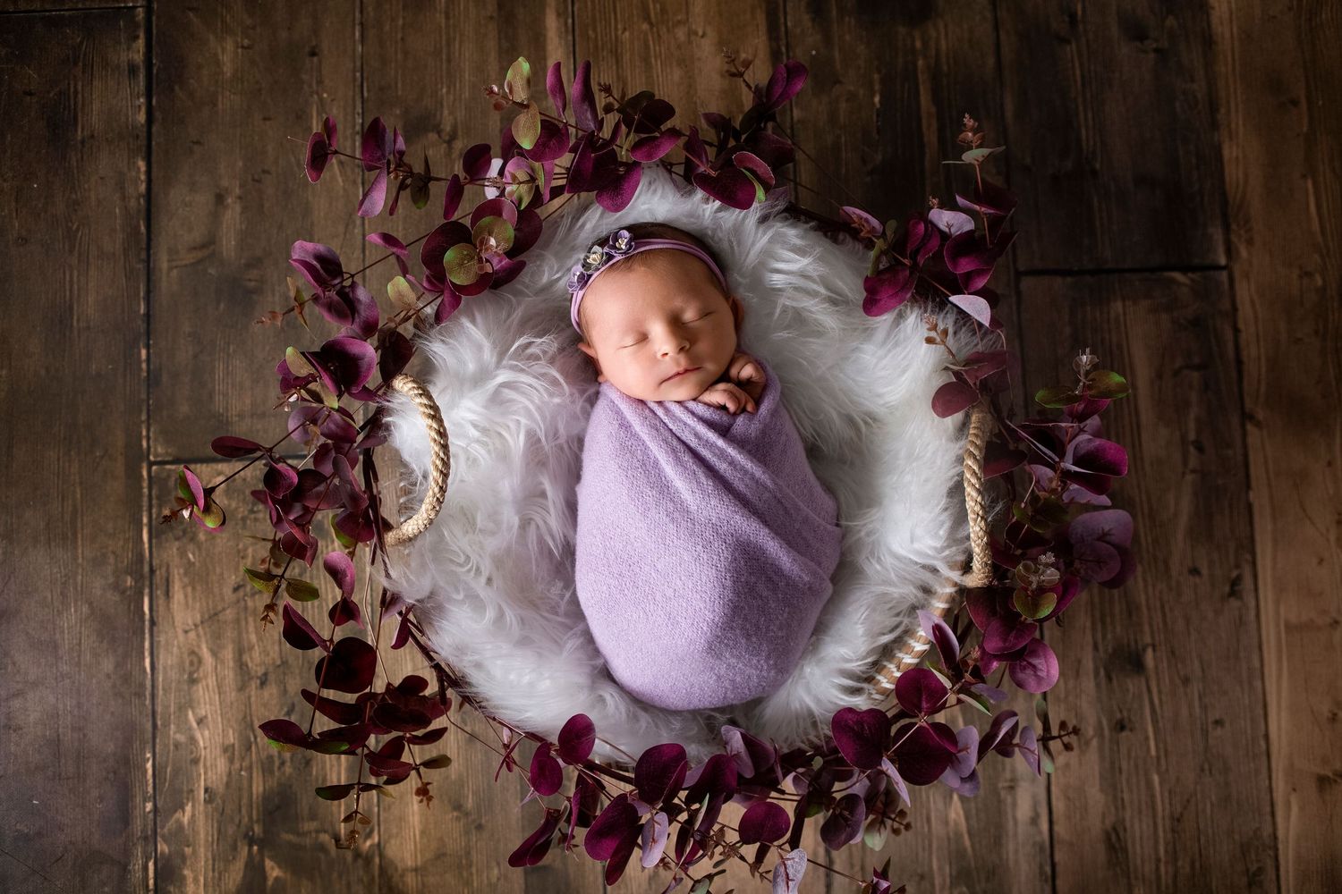 Newborn baby swaddled in purple wrap sleeping in a basket surrounded by purple floral decorations.