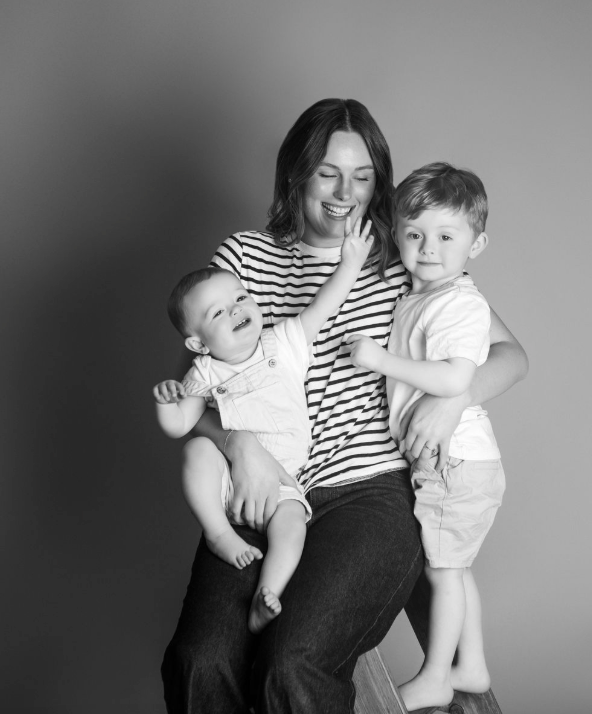 Black and white studio portrait of a smiling mother sitting with her baby and young toddler son.
