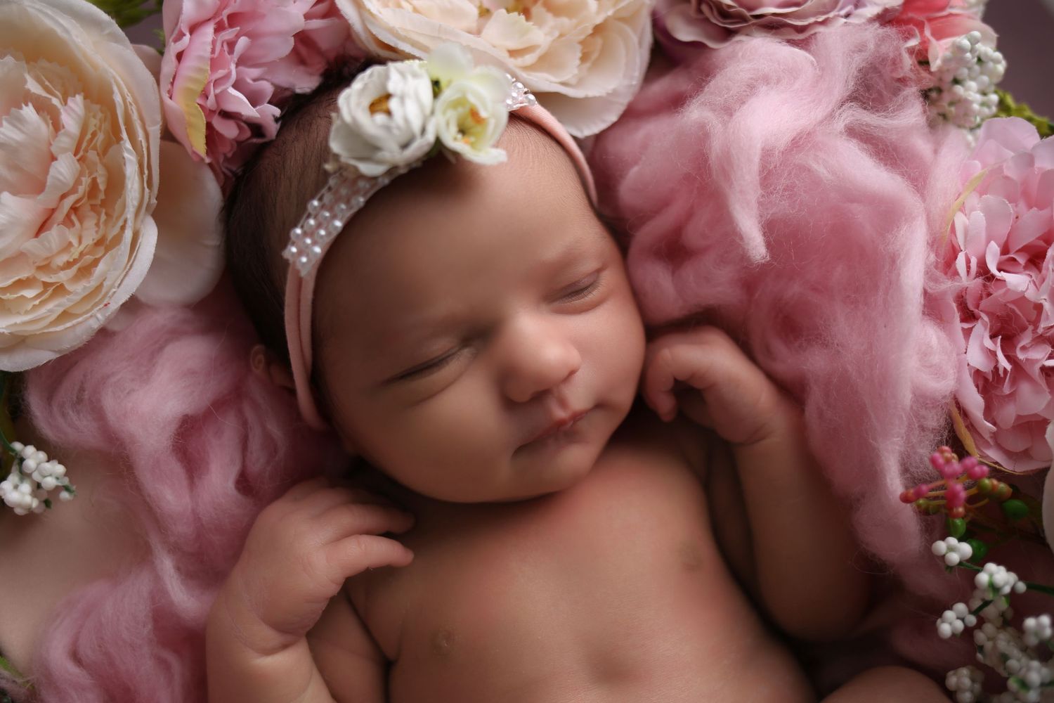 Newborn baby girl sleeping peacefully on pink floral backdrop with roses and a delicate flower headband.