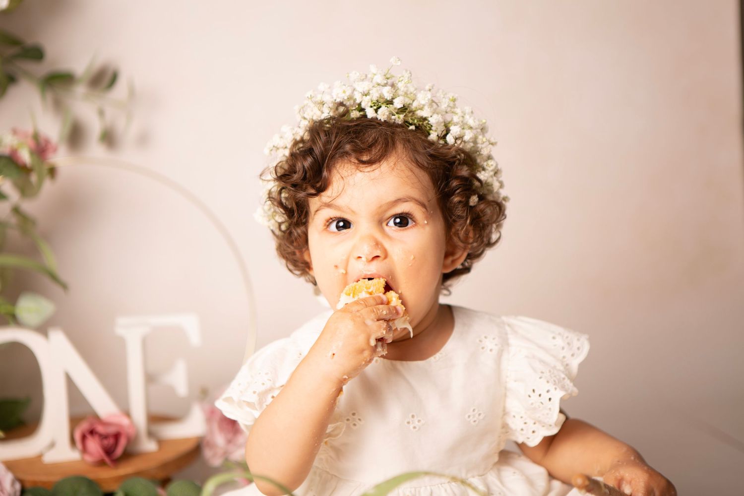 Toddler girl in white dress and flower crown eating cake during a first birthday cake smash photo session.