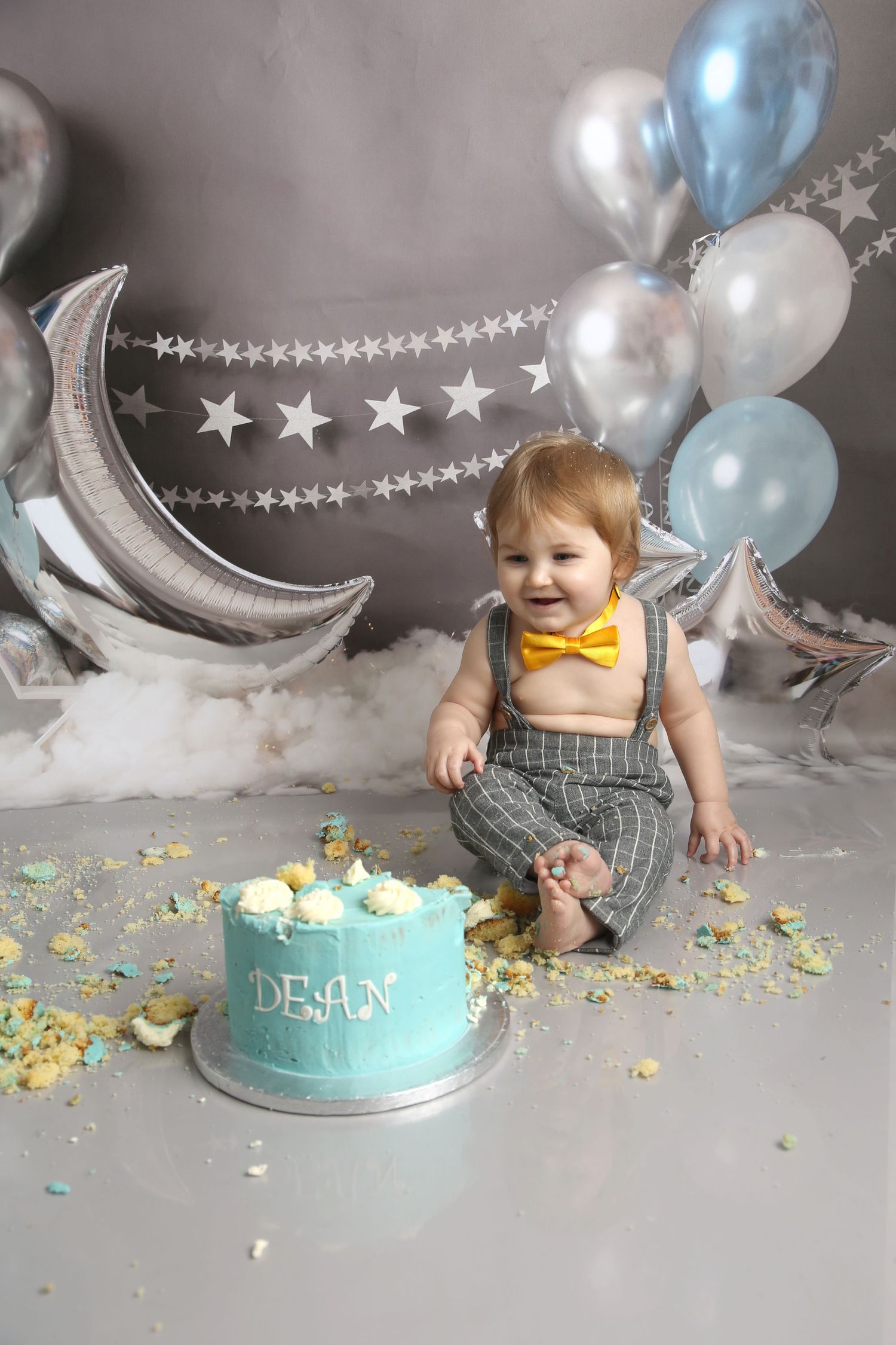 Baby boy Dean celebrates first birthday smashing teal cake, wearing gray suspenders and yellow bow tie, surrounded by silver balloons.