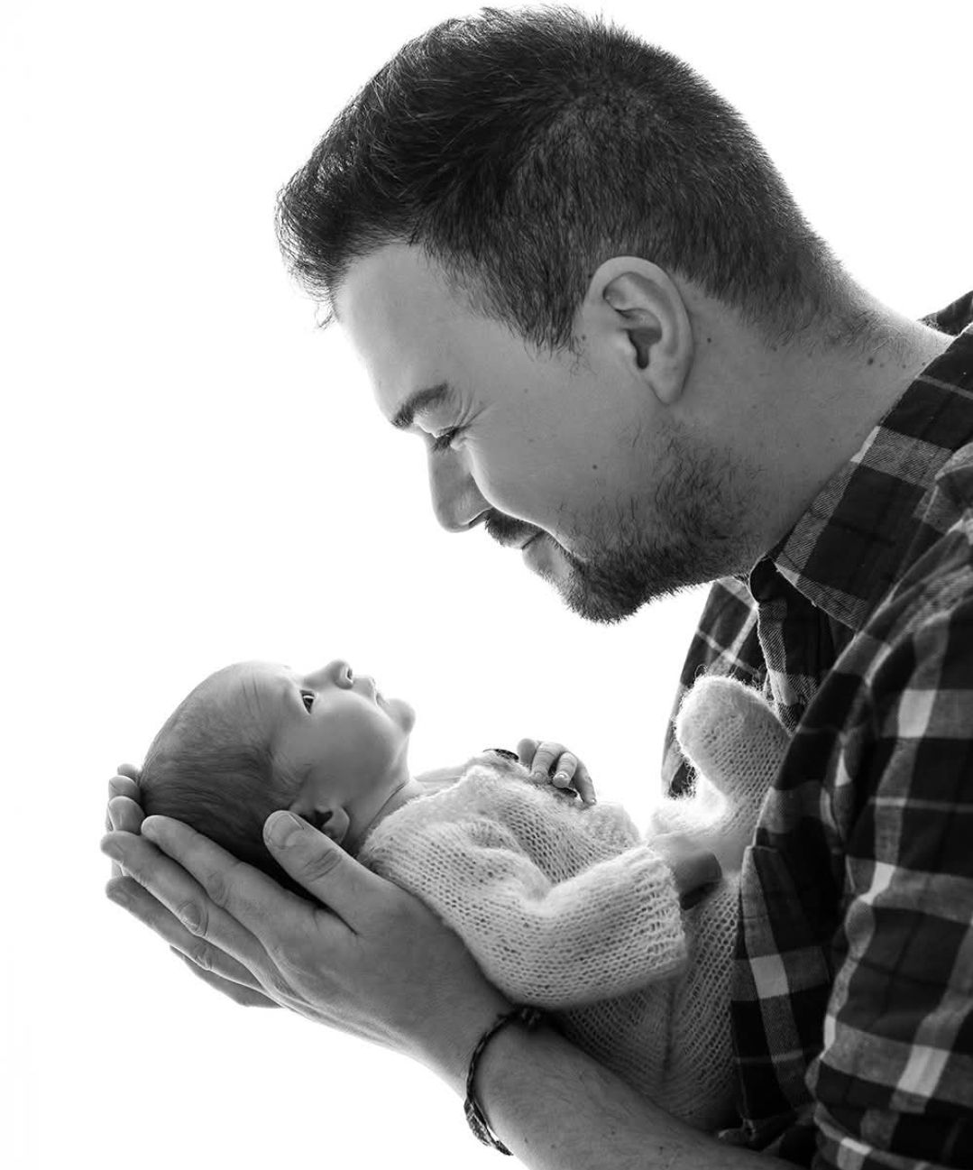 Black and white photo of a bearded father tenderly holding his newborn baby in both hands, gazing lovingly.