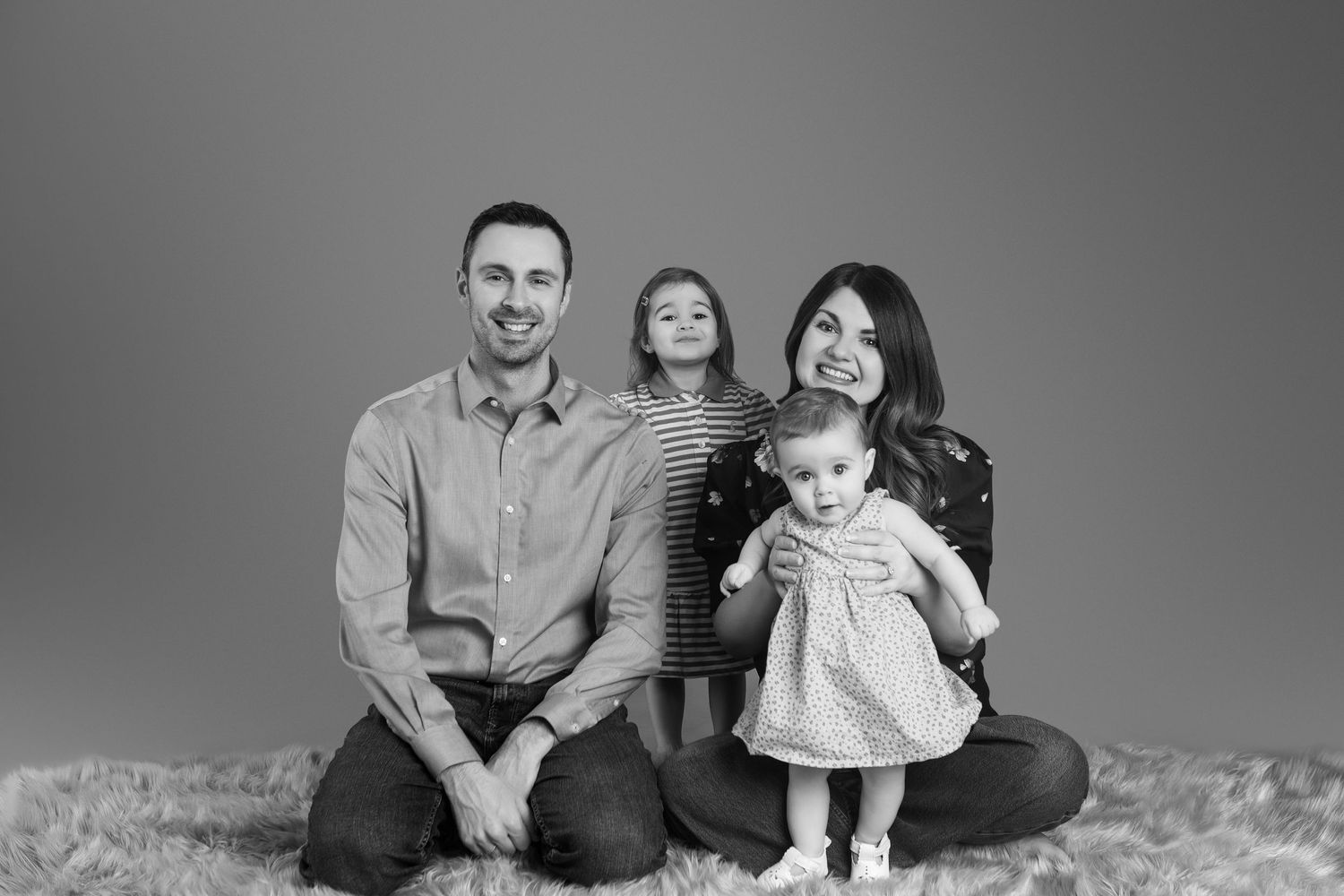 Black and white family portrait of parents with two young children sitting on a fluffy rug against a gray backdrop.