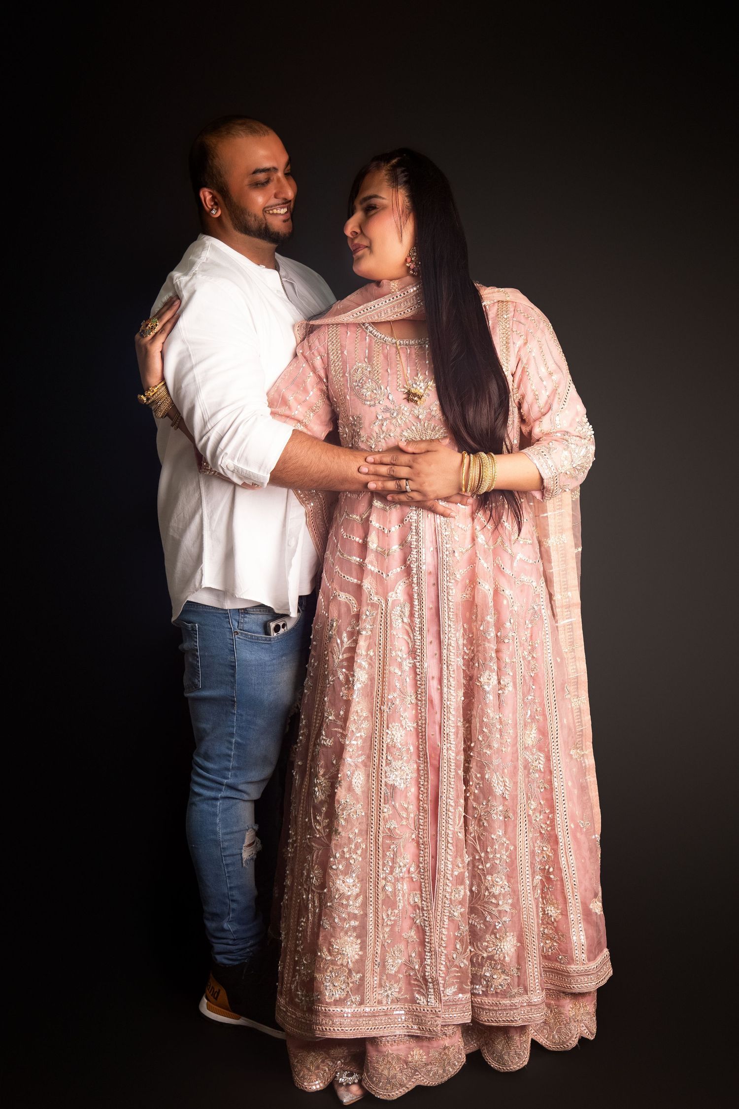 Couple in studio portrait, man in white shirt embracing woman in elegant pink embroidered anarkali gown.