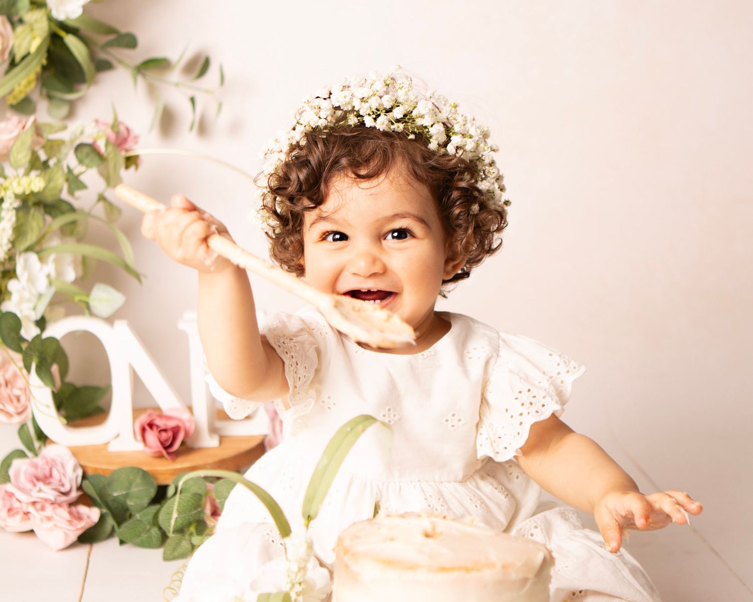 Joyful toddler girl in white dress and floral crown smashing a cake, surrounded by pink roses.