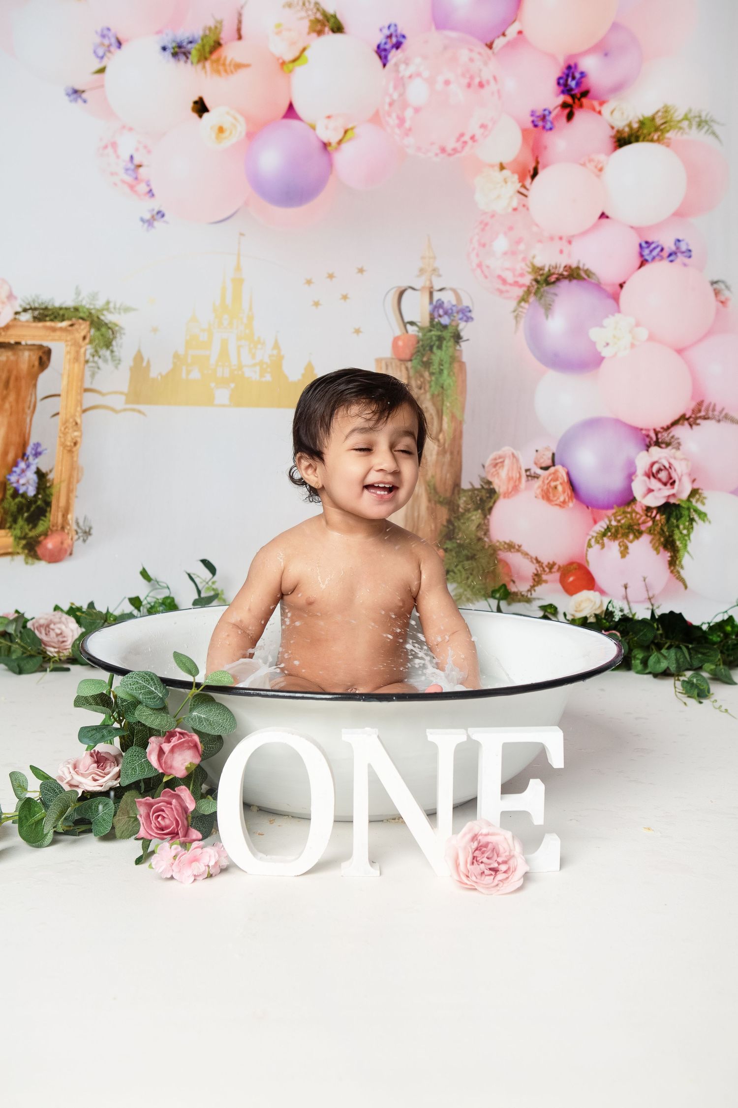 Baby smiling in a tub at a princess-themed first birthday cake smash with pink balloons and ONE sign.