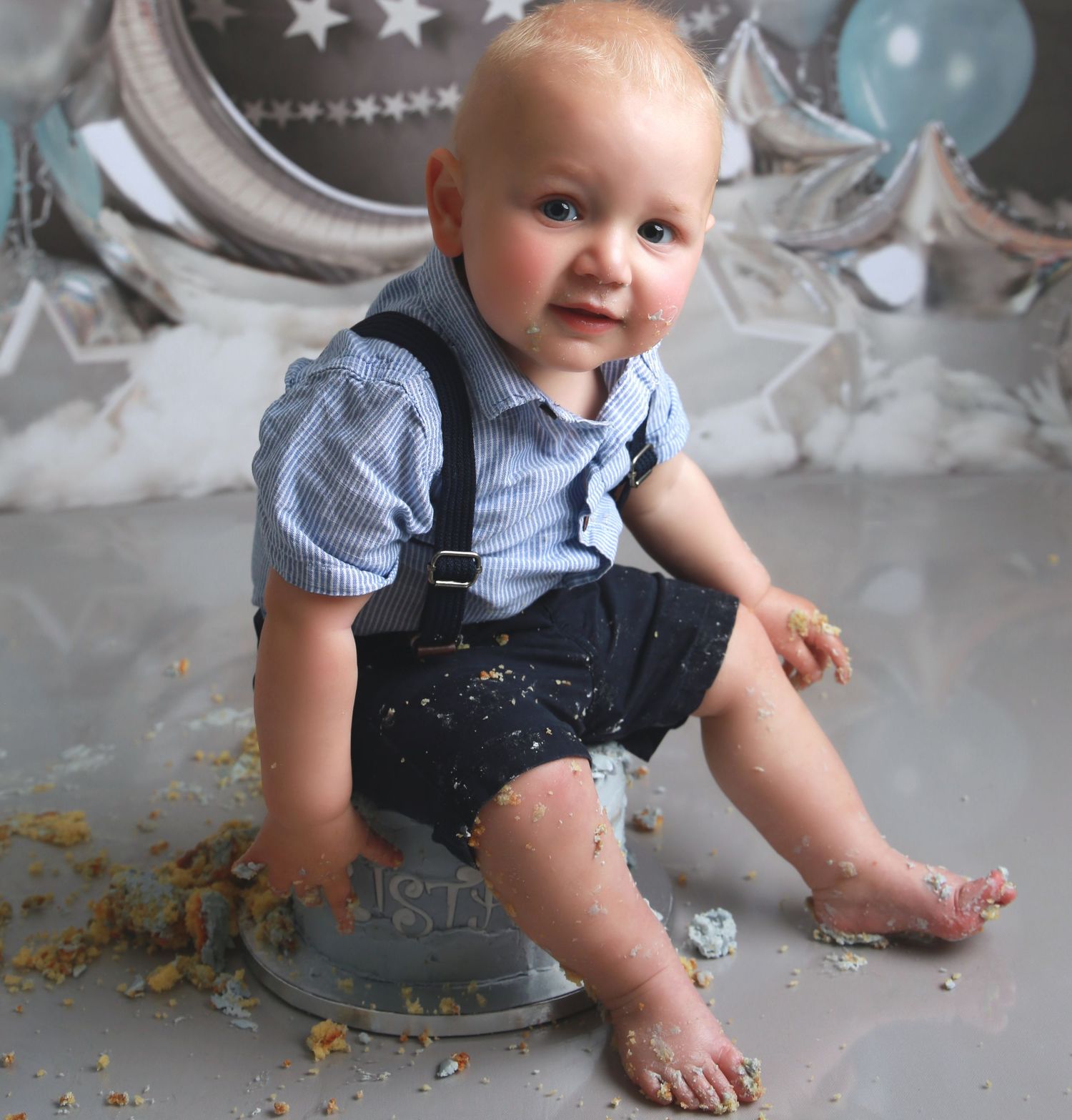 Baby boy in suspenders and blue shirt sits on a tin smashing his first birthday cake on a gray backdrop.