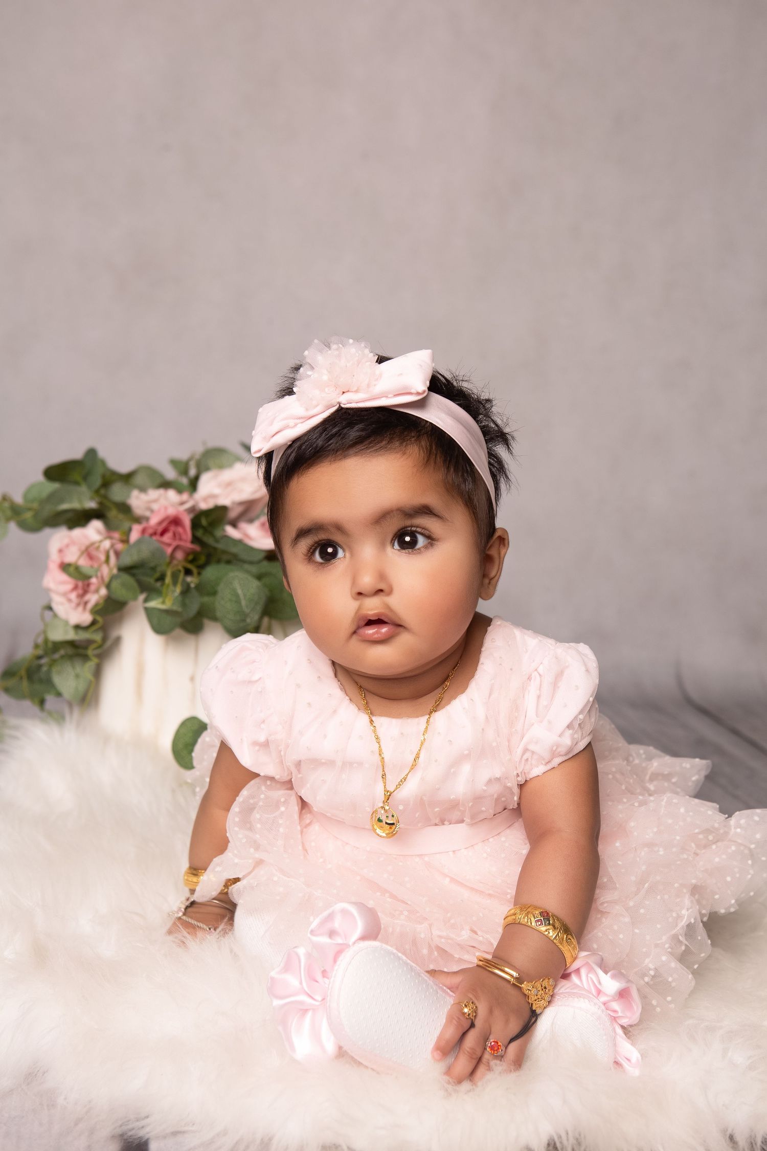 Baby girl in pink dress and headbow with gold jewelry sits on white fur rug with roses backdrop.