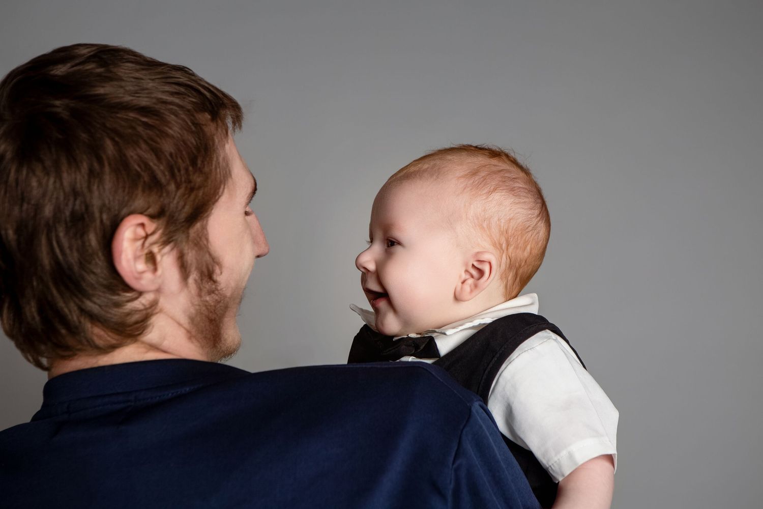 Father holding smiling baby in black vest during professional newborn portrait session on gray background.
