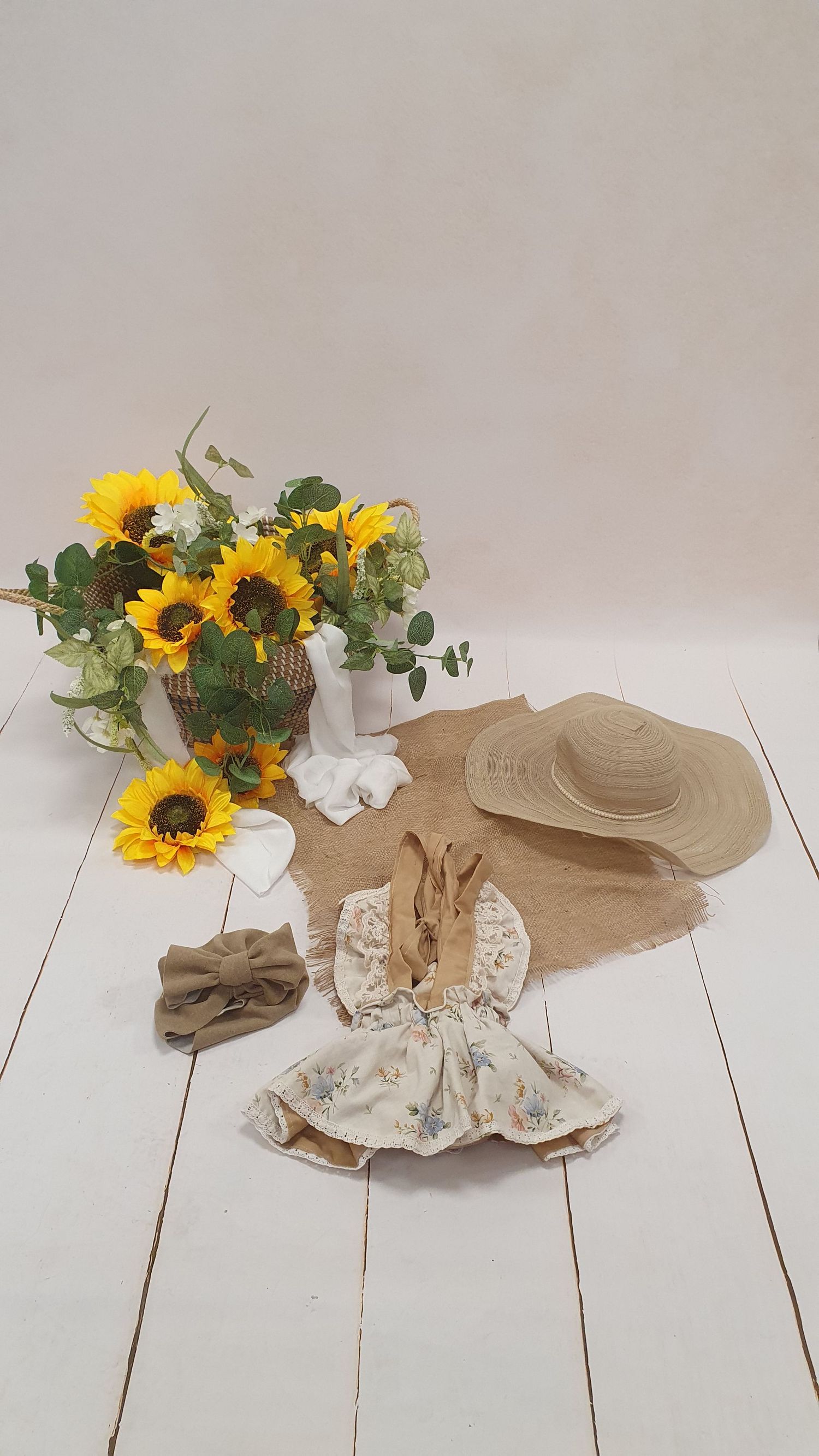 Floral baby dress, straw hat, bow headband, and sunflowers arranged on a rustic white wood surface.