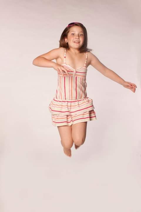 A young girl in a striped sundress joyfully jumping in the air against a light background.