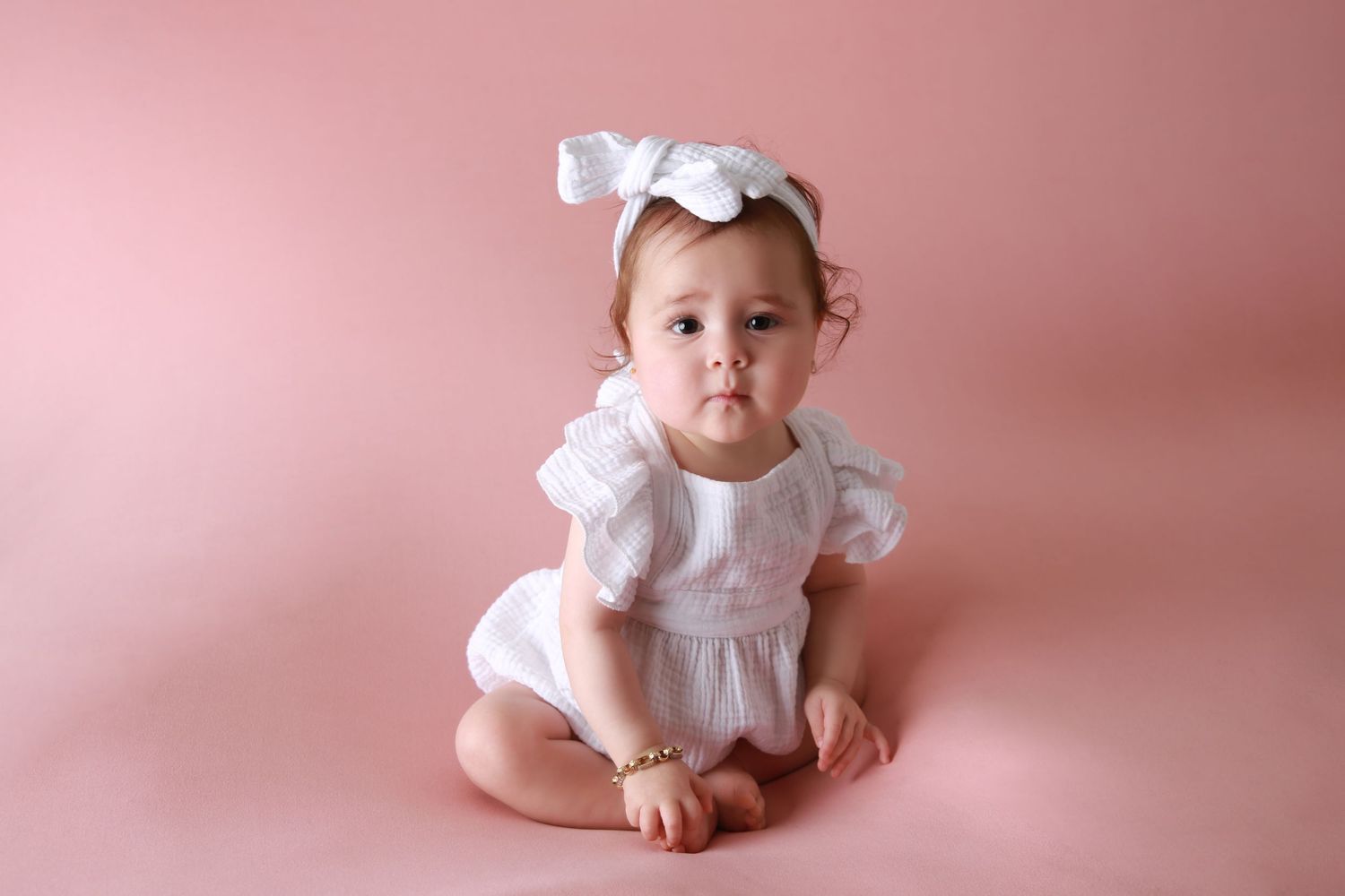 Adorable baby girl in white ruffled romper and bow headband sitting on pink background.