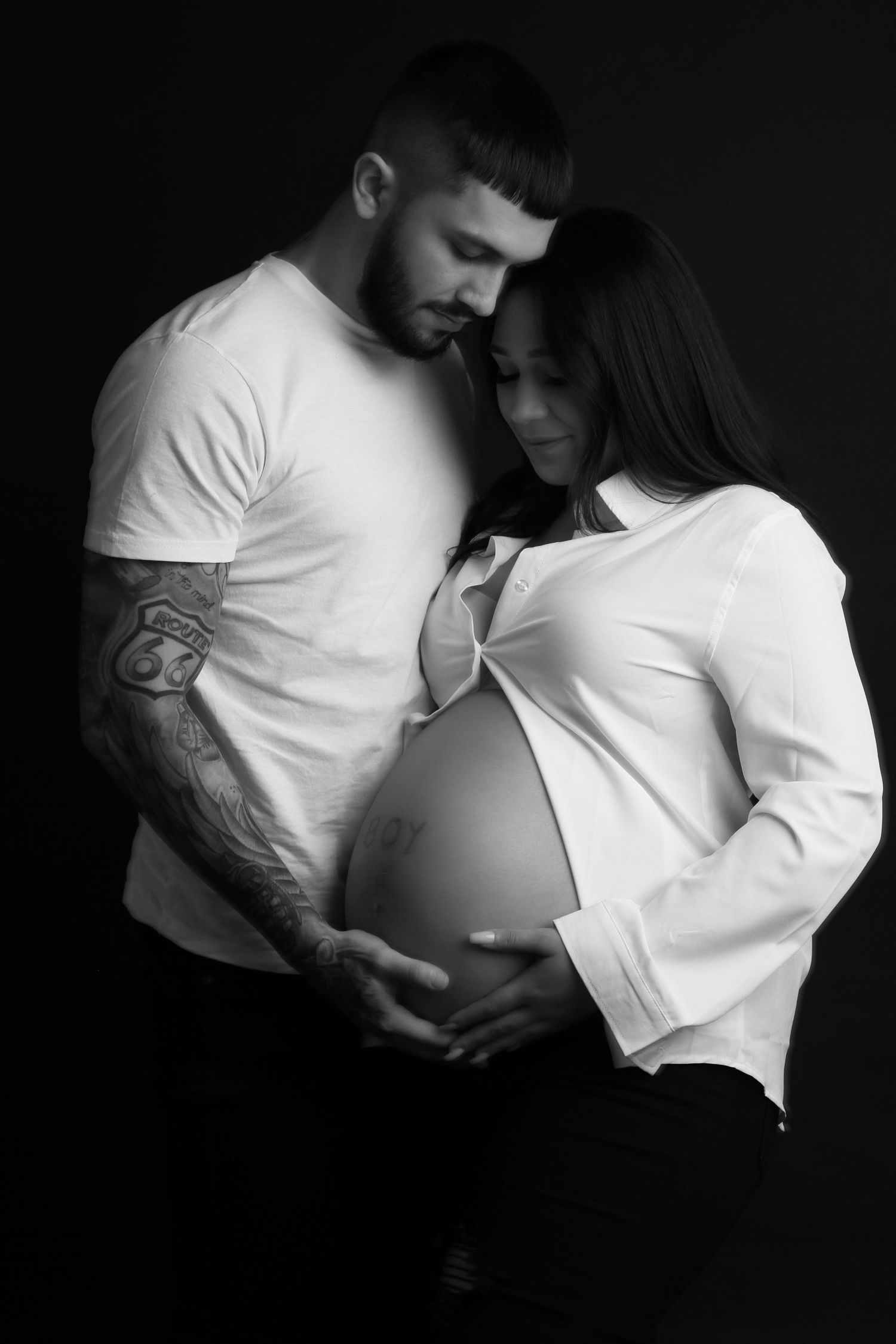 Black and white maternity photo of tattooed man embracing pregnant woman in white shirt.